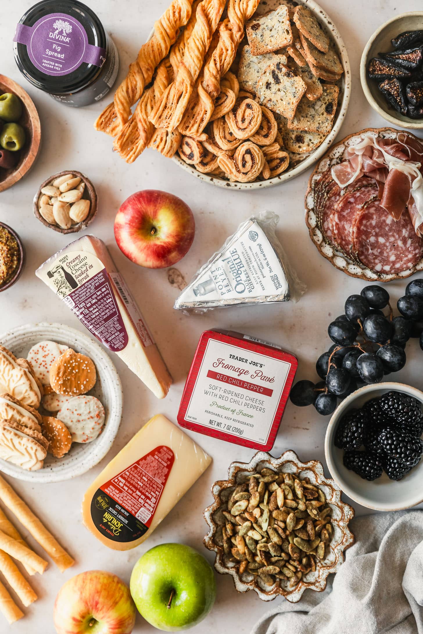Cheeses, fall fruits, apples cookies, a jar of fig jam, and brown and white plates of crackers, olives, meats, cookies, and pepitas on a beige counter.