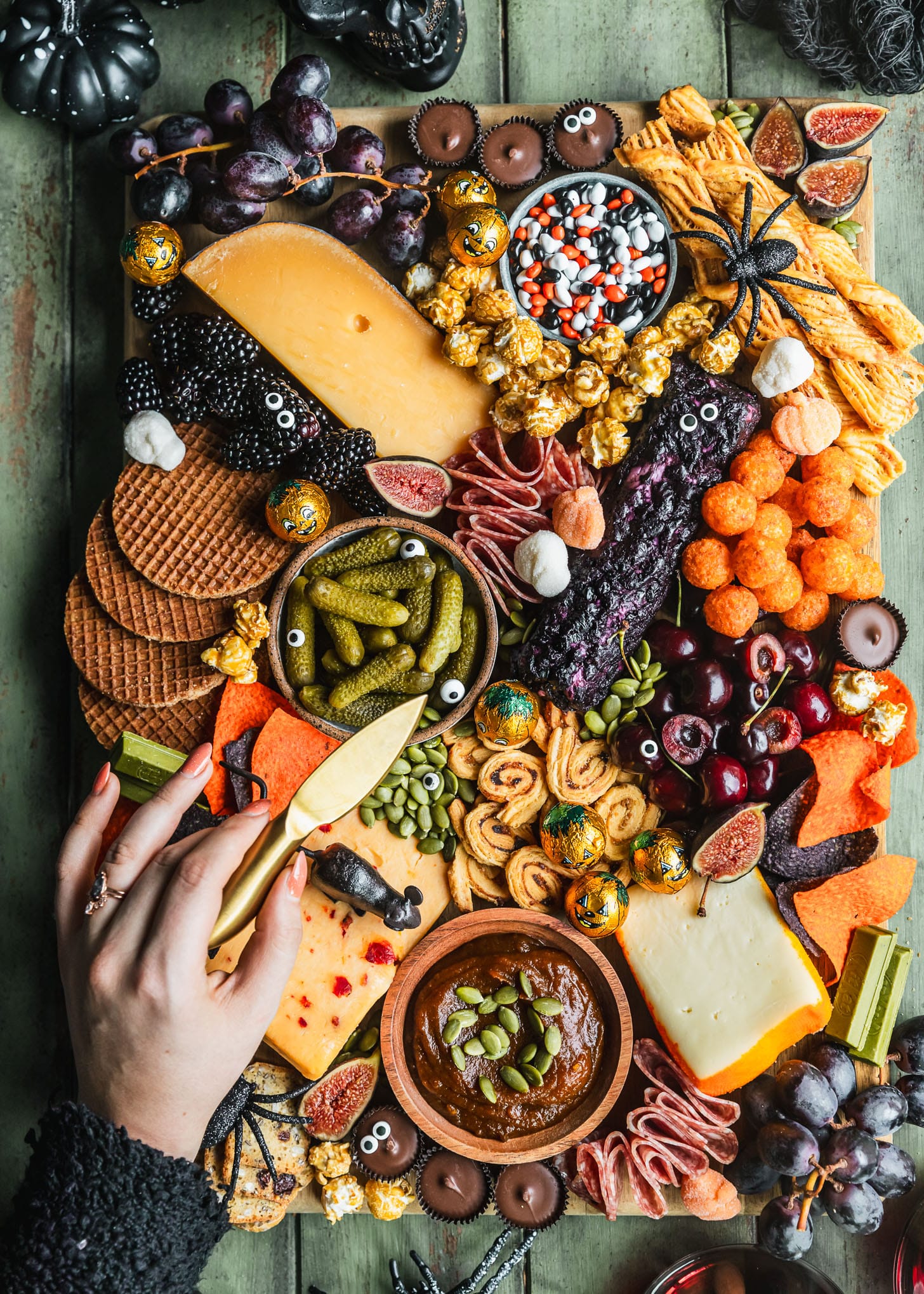 A woman's hand holding a gold cheese knife and reaching for cheese on a Halloween charcuterie board next to a black skeleton and glasses of red wine on a wood green board.