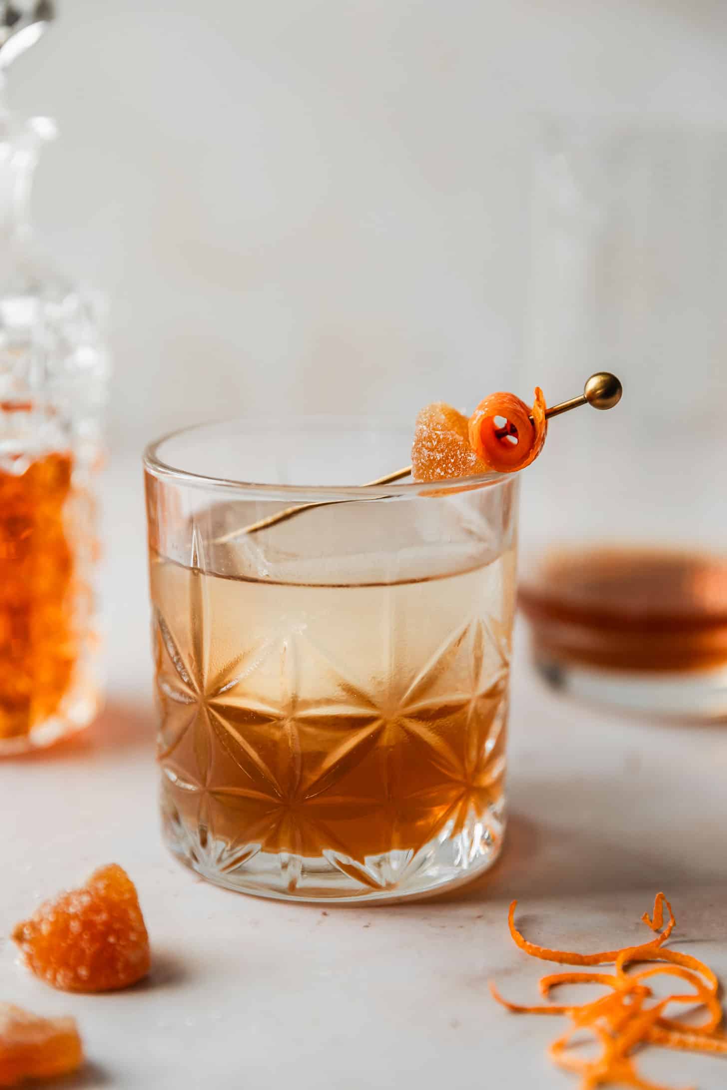 A ginger old fashioned on a tan marble counter next to a decanter of bourbon, cocktail mixing glass, crystallized ginger, and orange peels.