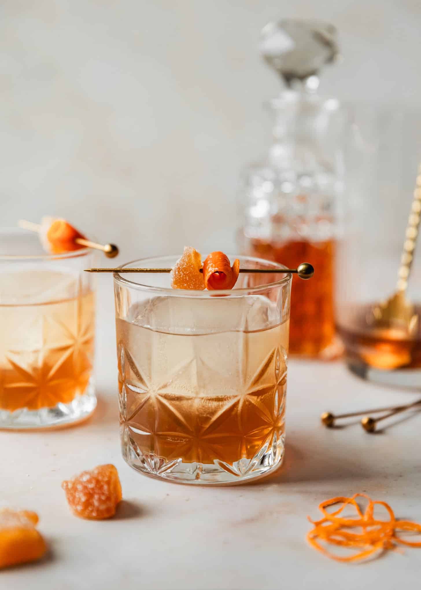Two ginger old fashioned cocktails on a beige counter next to a cocktail mixer, decanter of bourbon, crystalized ginger, and orange peels.