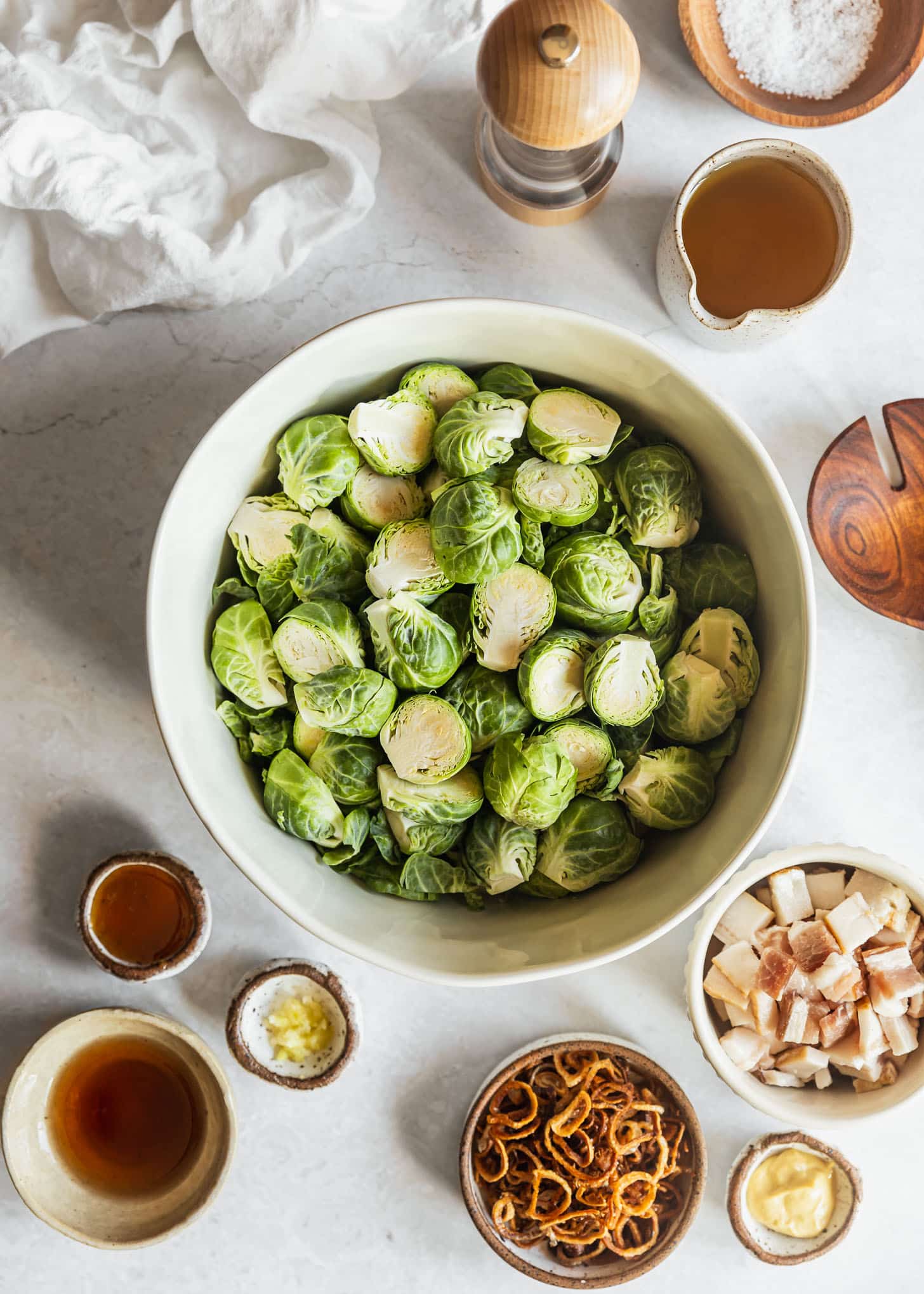 White bowls of Brussels sprouts, raw bacon, fried shallots, Dijon, honey garlic, vinegar, and oil on a white marble counter next to a white linen and wood spoon.