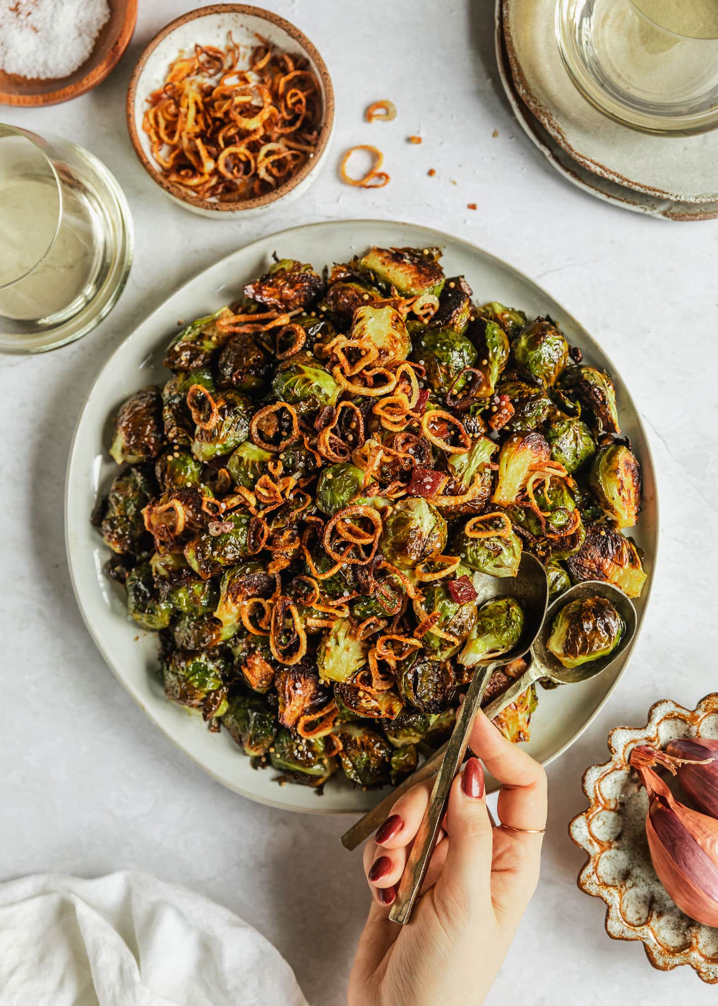 A hand spooning crispy Brussels sprouts off of a white platter next to two glasses of white wine and brown and white bowls of fried shallots and raw shallots on a marble counter.