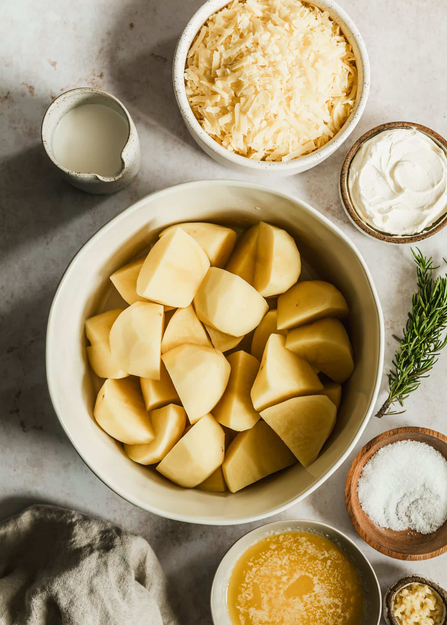 White and brown bowls of white cheddar, potatoes, creme fraiche, cream, salt, melted butter, and garlic on a beige table next to rosemary and a tan linen.