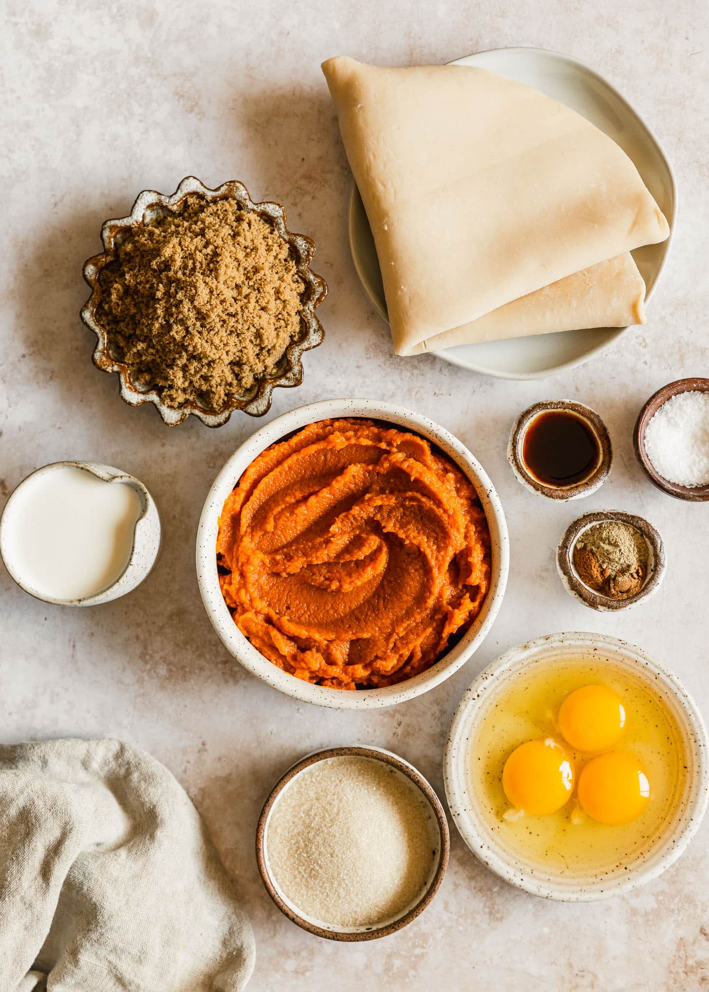 White and brown bowls of pie dough, brown sugar, pumpkin, half and half, spices, vanilla, salt, eggs, and sugar on a beige counter.