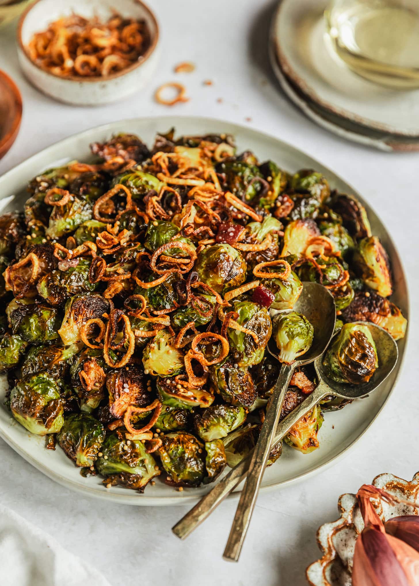 A white platter of caramelized Brussels sprouts with bacon on a white marble counter next to a white bowl of fried shallots, grey plates, and a white bowl of shallots.
