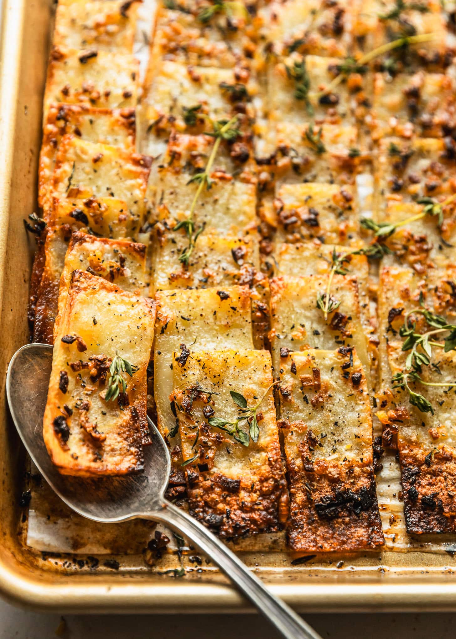 A spoon scooping domino potatoes with shallots, rosemary, and thyme off of a gold sheet pan.