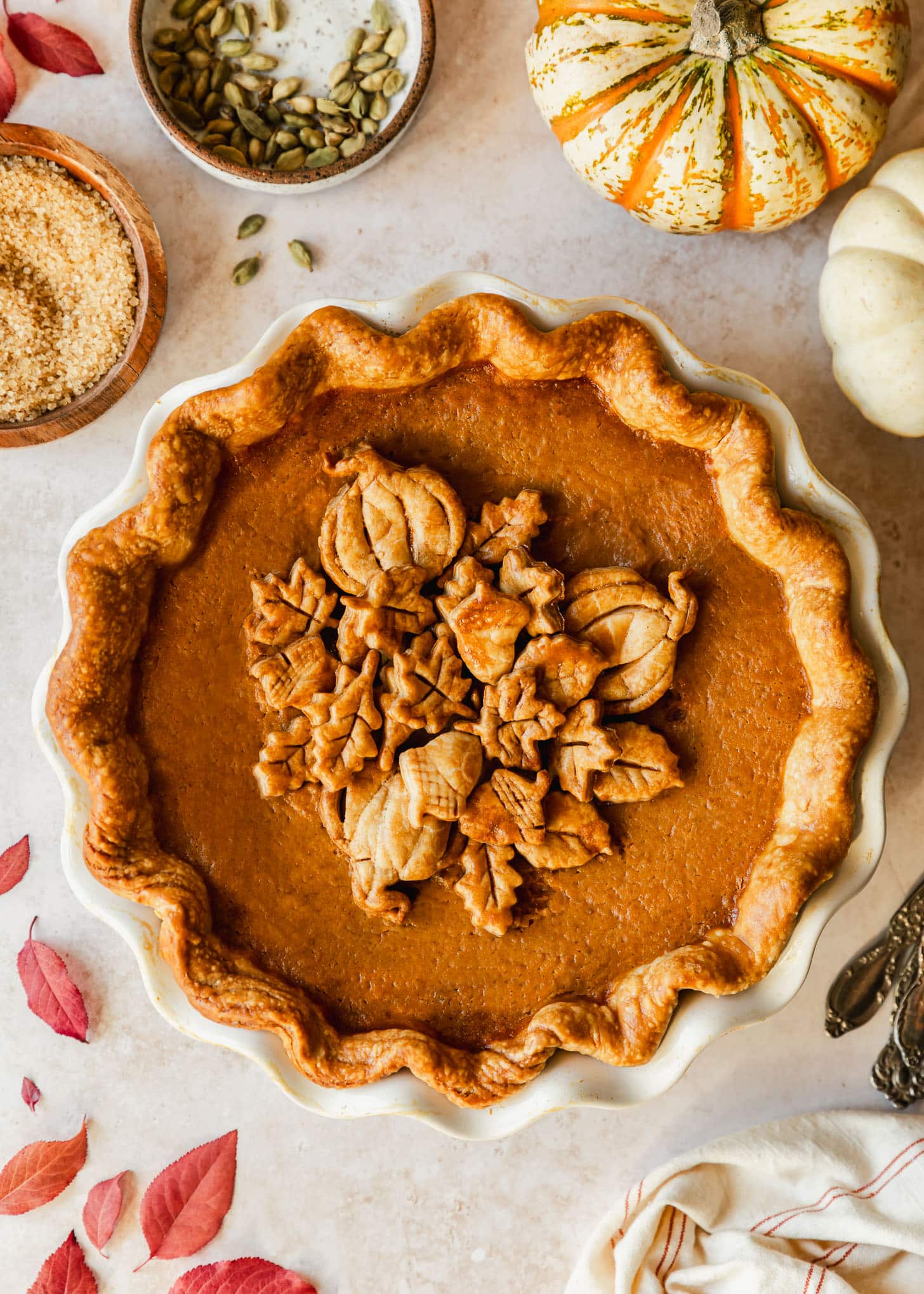 A white pie plate with cardamom pumpkin pie on a beige counter next to a white and orange pumpkin, white pumpkin, white napkin, red leaves, and brown bowls of sugar and cardamom pods.