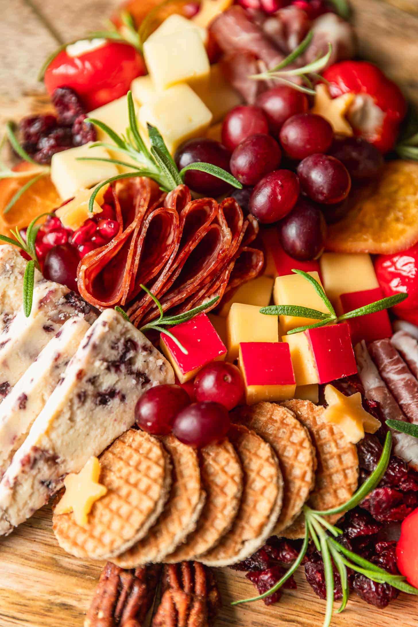 A close up of cheeses, meats, grapes, peppers, rosemary, cookies, and dried fruit on a wood platter.