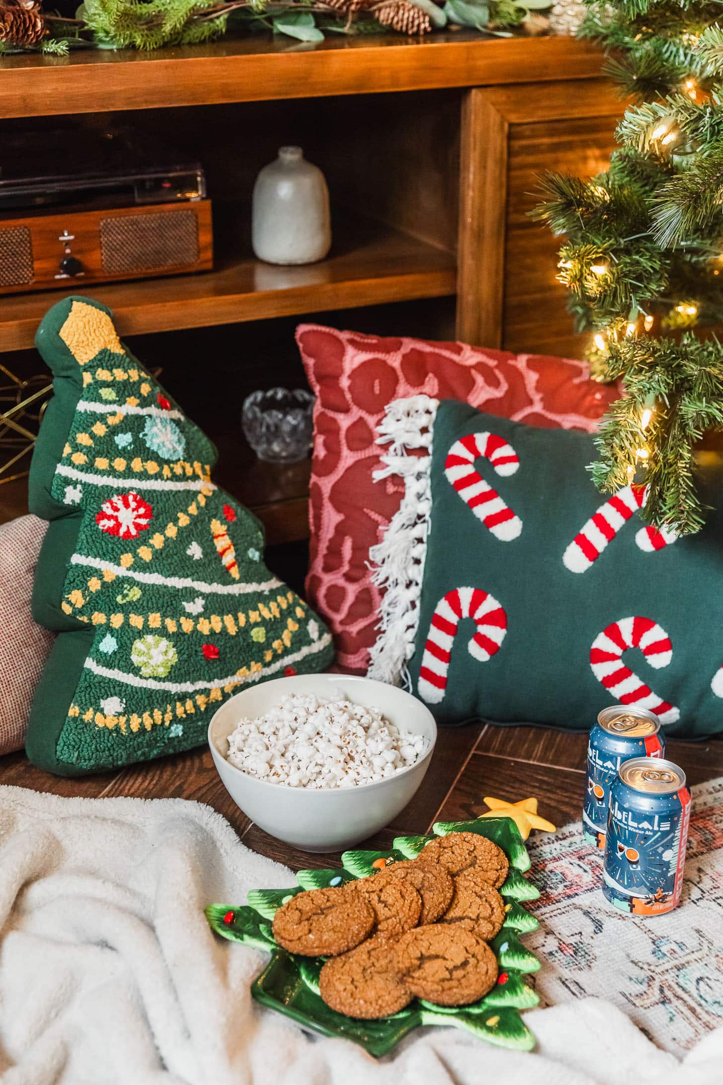 A Christmas tree plate with cookies on a white blanket next to a white bowl of popcorn, cans of beer, and Christmas pillows in front of a wood media console.
