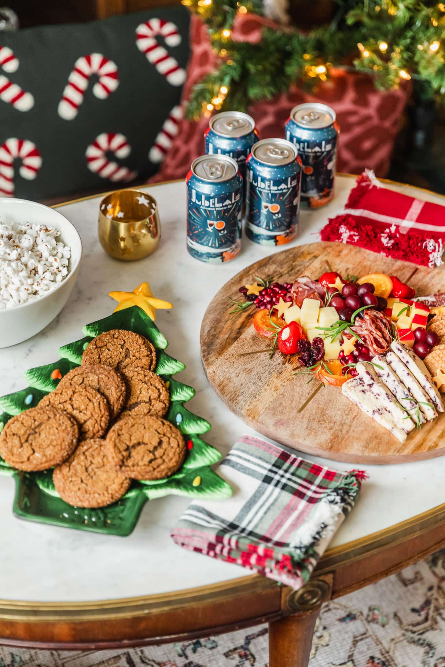 A Christmas movie night party with a charcuterie board, white bowl of popcorn, beer, and Christmas tree plate of cookies on a marble coffee table next to a Christmas tree.