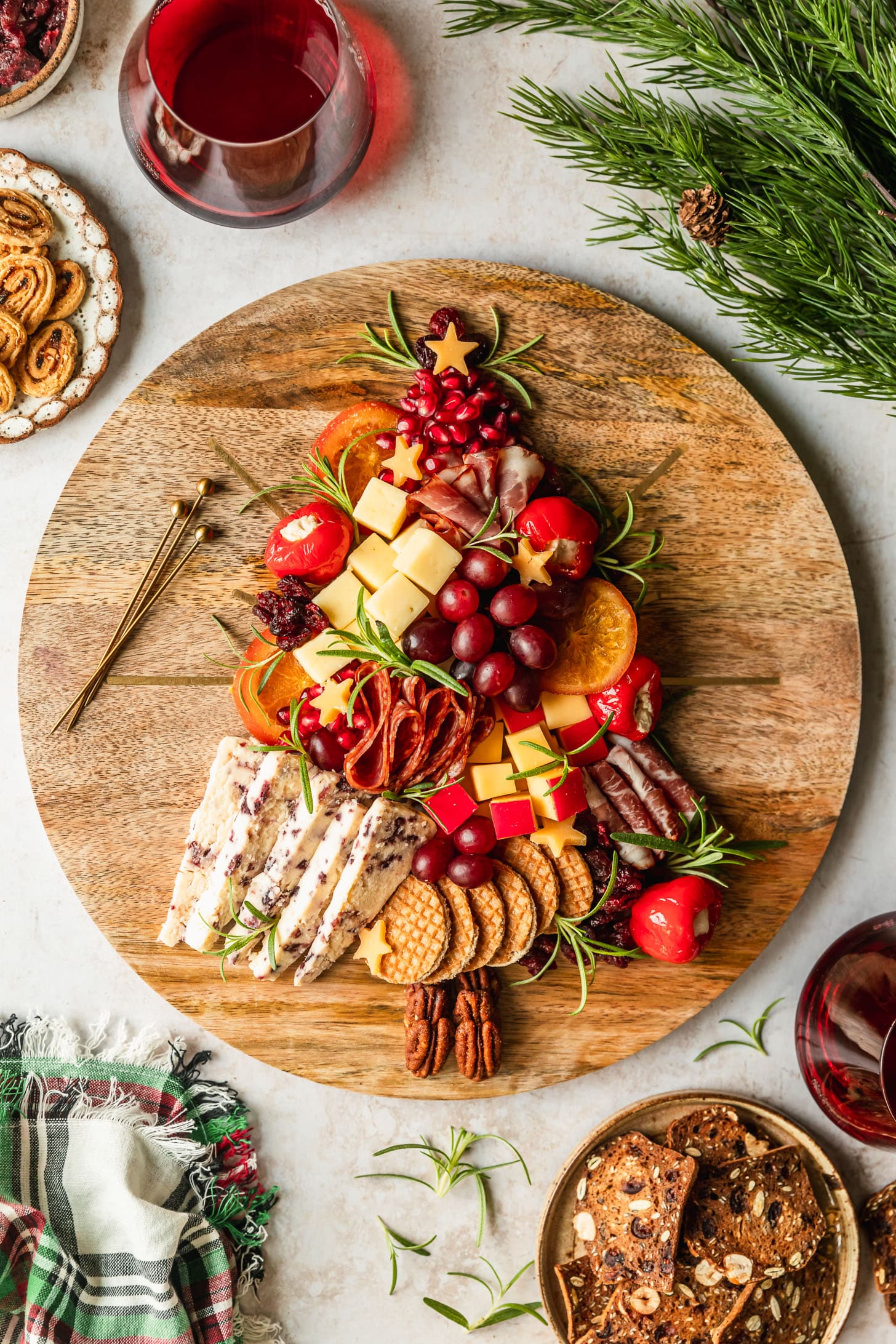 A Christmas tree charcuterie board on a wood board next to a plaid linen, glasses of red wine, and brown plates of crackers.