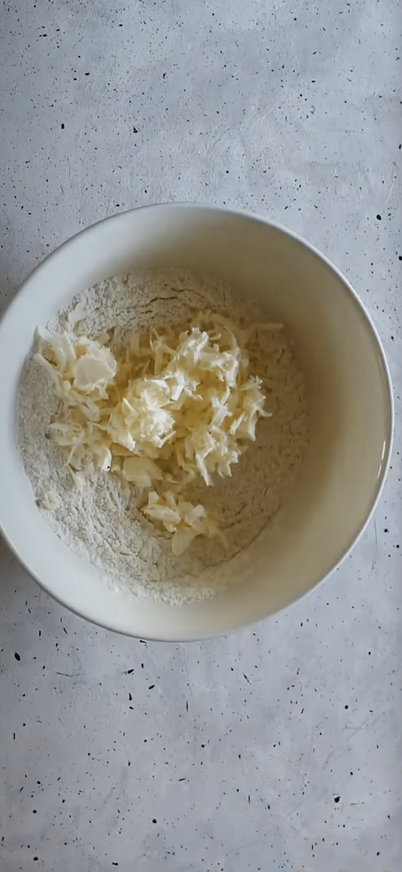 A white bowl of flour and grated butter on a grey counter.