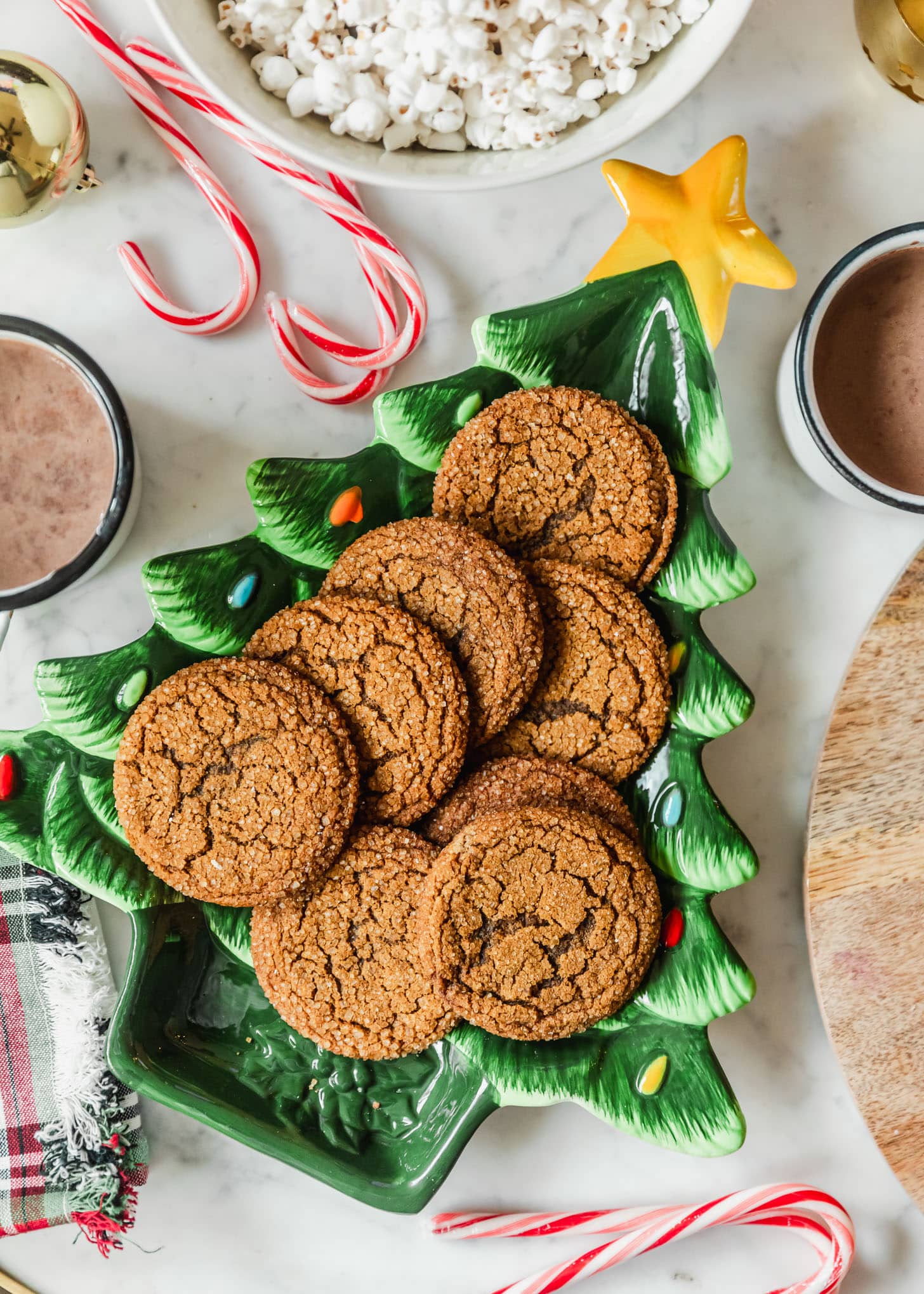 A Christmas movie night party. A Christmas tree plate with cookies on a marble coffee table next to candy canes, a bowl of popcorn, mugs of hot chocolate, and plaid linens.