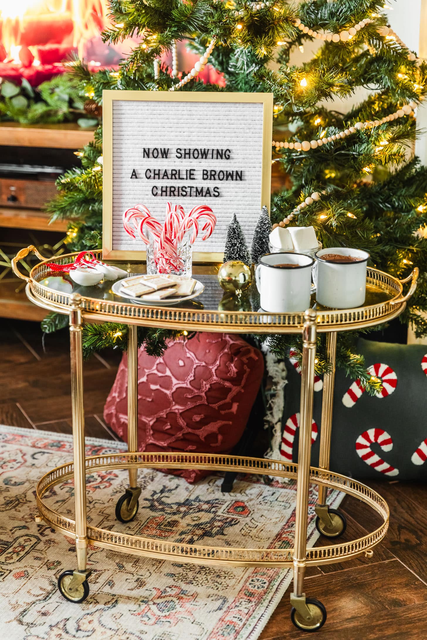 A Christmas movie night party. A gold bar cart topped with white mugs of hot chocolate, a letter board sign, candy canes, chocolate, and marshmallows is placed in front of a Christmas tree.