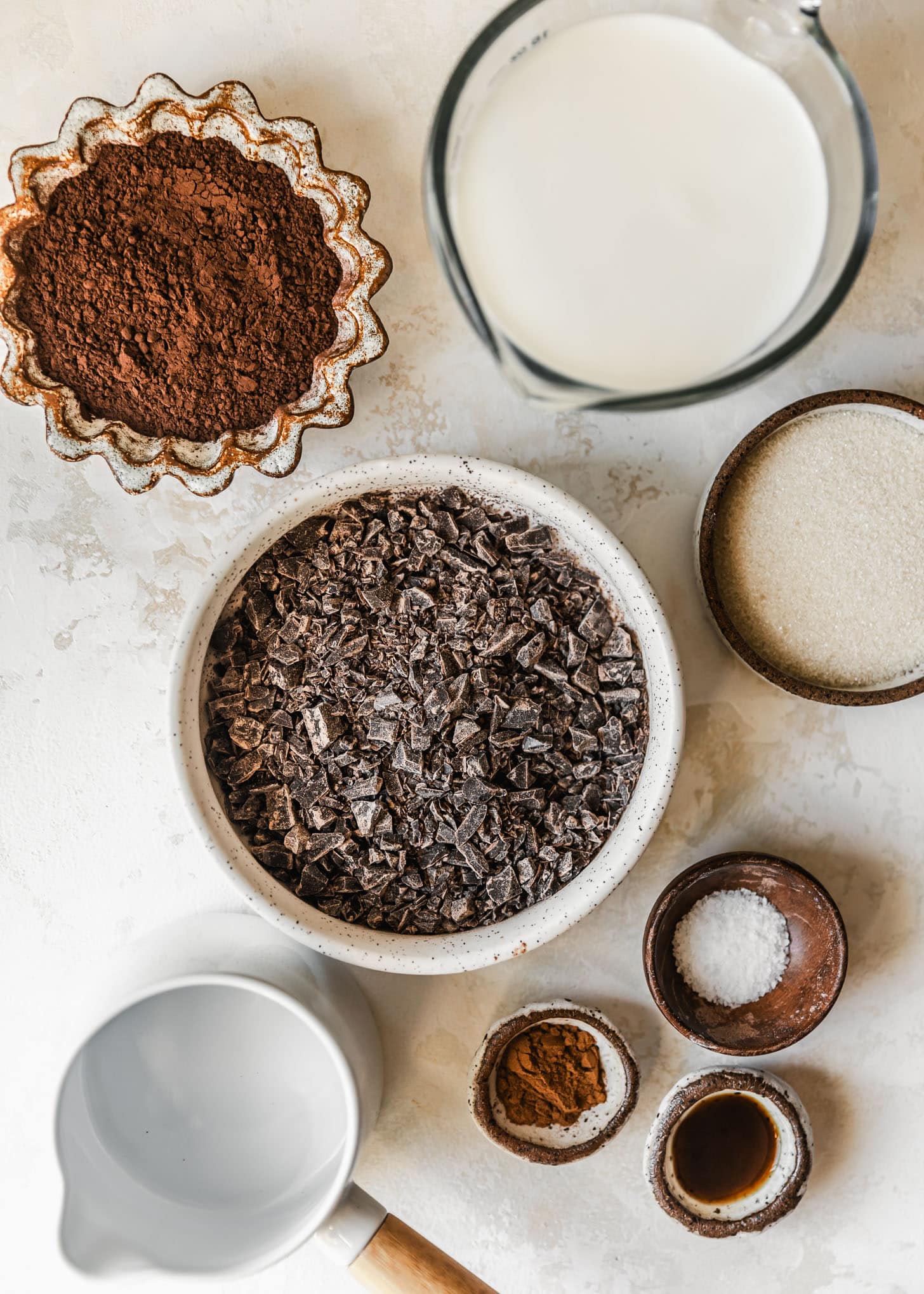 White and brown bowls of cocoa powder, milk, semisweet chocolate, sugar, salt, cinnamon, and vanilla on a beige counter.