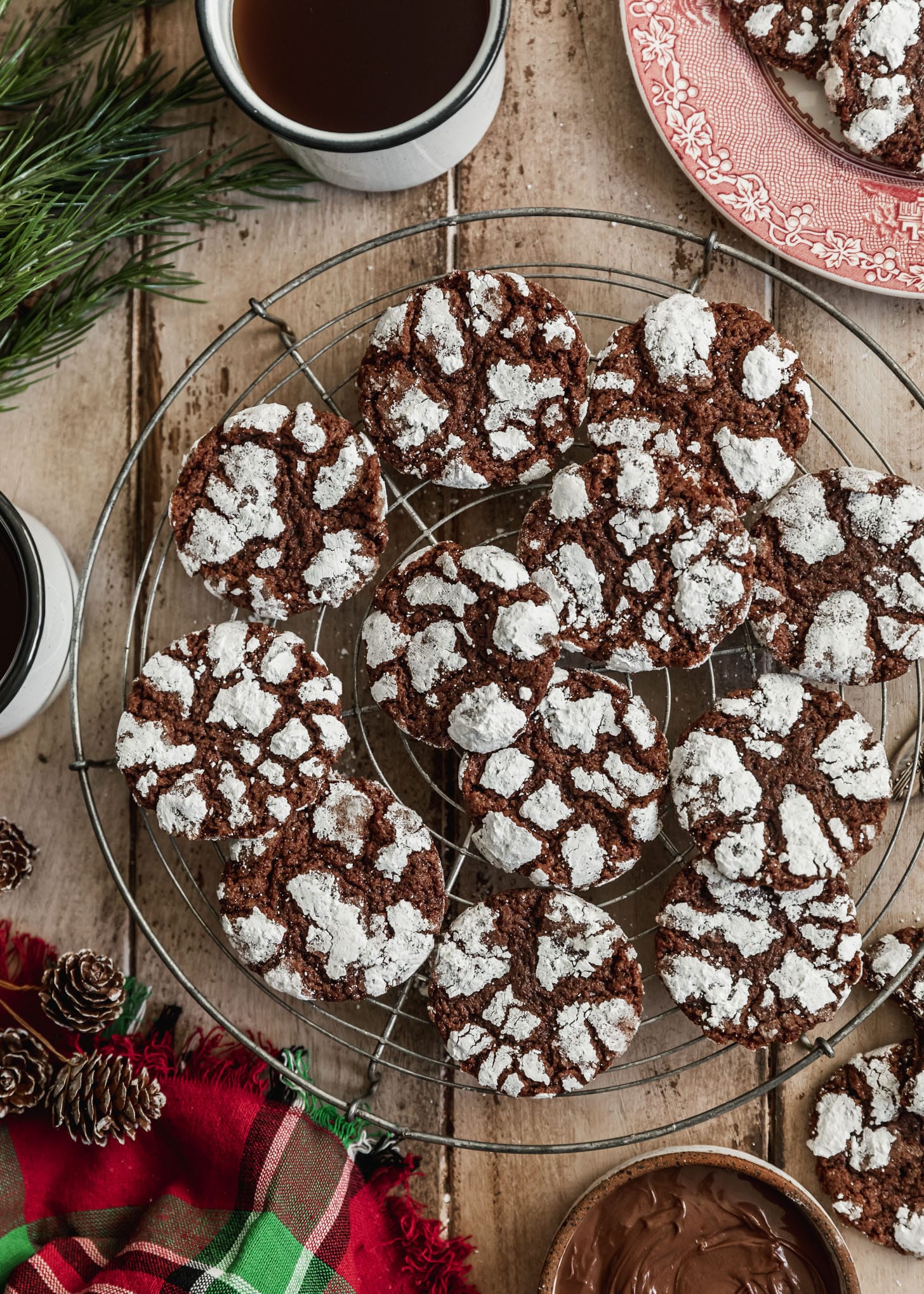 A wire rack of mocha crinkle cookies on a wood table next to a red plaid linen, pinecones, a white bowl of Nutella, and white mugs of coffee.
