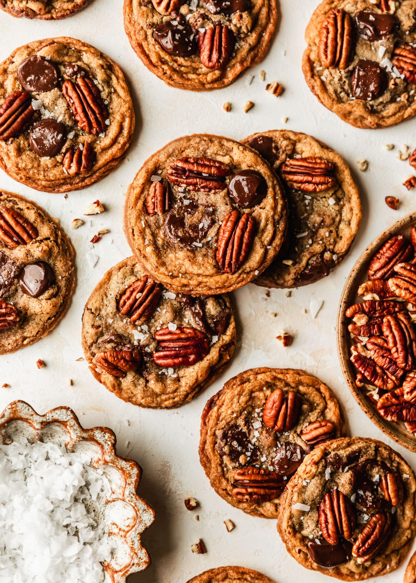 Piles of pecan chocolate chip cookies with brown butter on a tan background next to brown bowls of pecans and flaky salt.