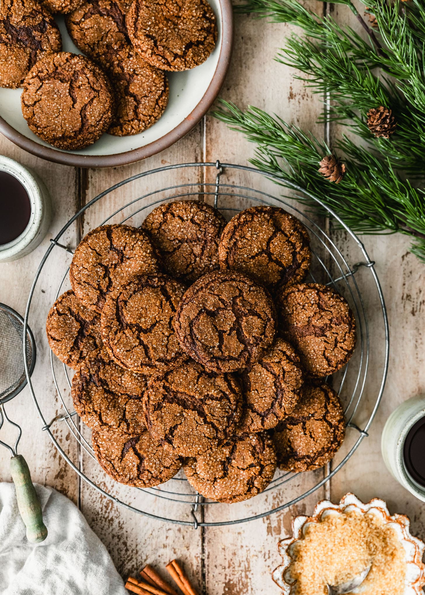 A wire rack of chewy molasses crinkle cookies on a wood table next to garland, grey cups of coffee, a white bowl of raw sugar, and a brown plate of cookies.
