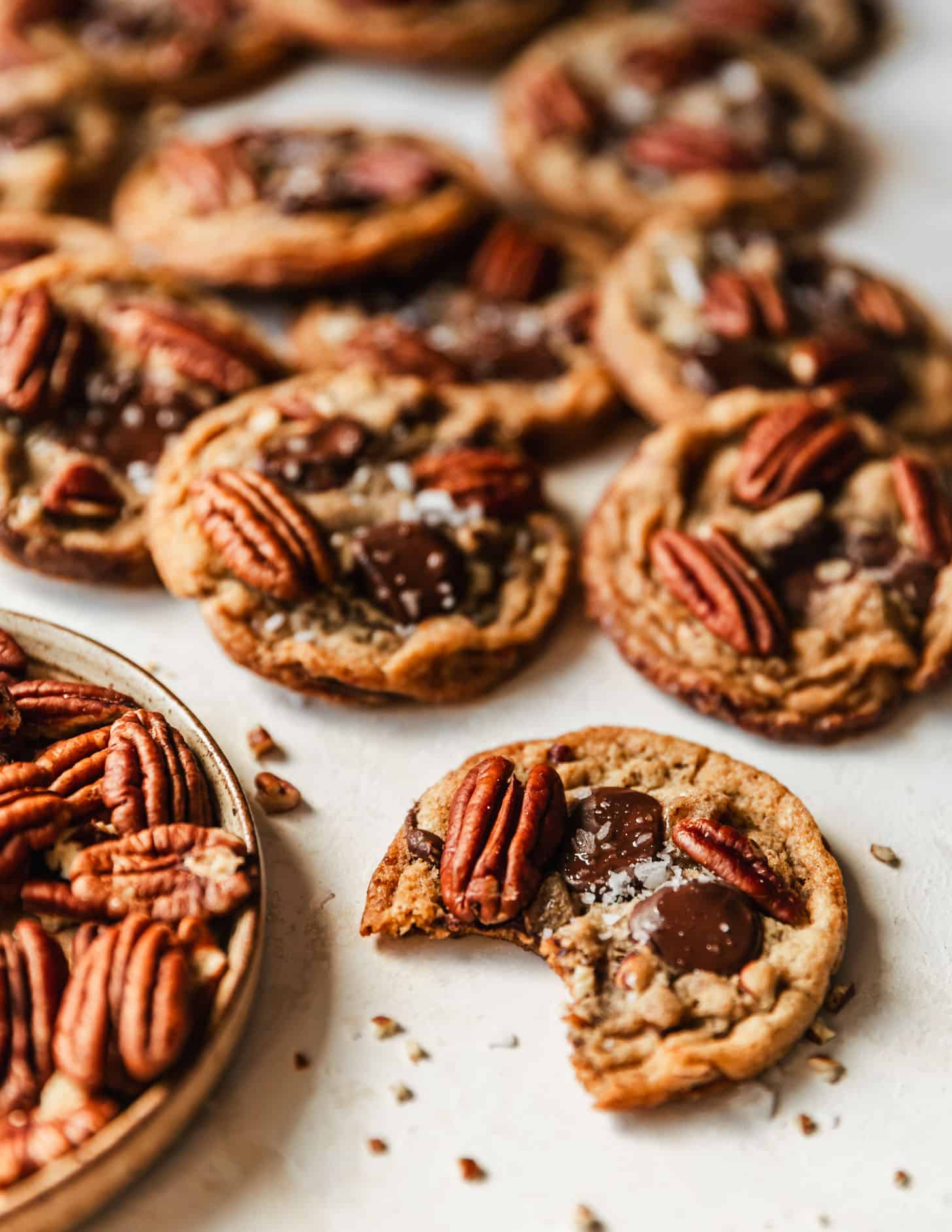 A pecan chocolate chip cookie with a bite taken out of it on a beige counter next to a brown plate of pecans and piles of cookies.