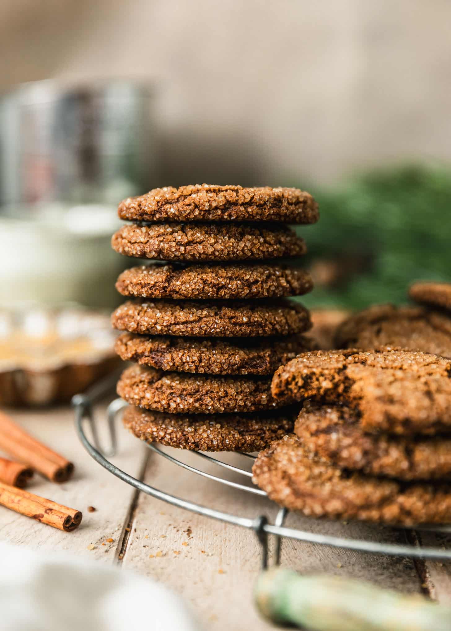 A stack of molasses crinkle cookies on a wire rack next to cinnamon sticks, garland, and a beige linen on a wood table.