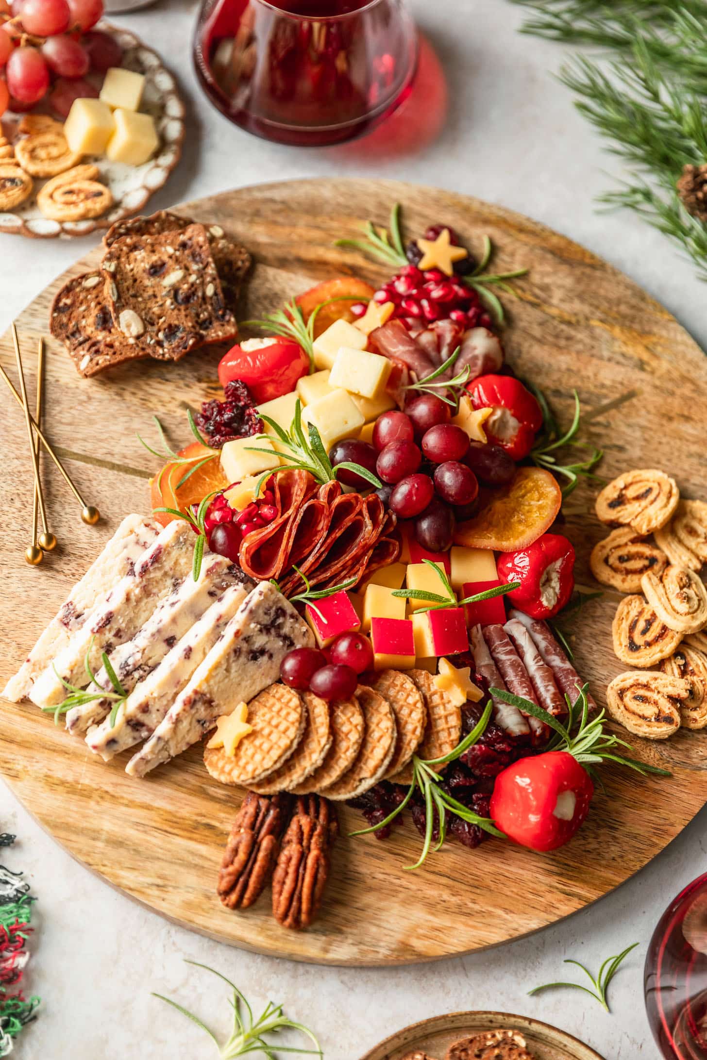 A Christmas tree charcuterie board on a tan counter next to glasses of red wine, garland, and white plates of crackers and fruit.