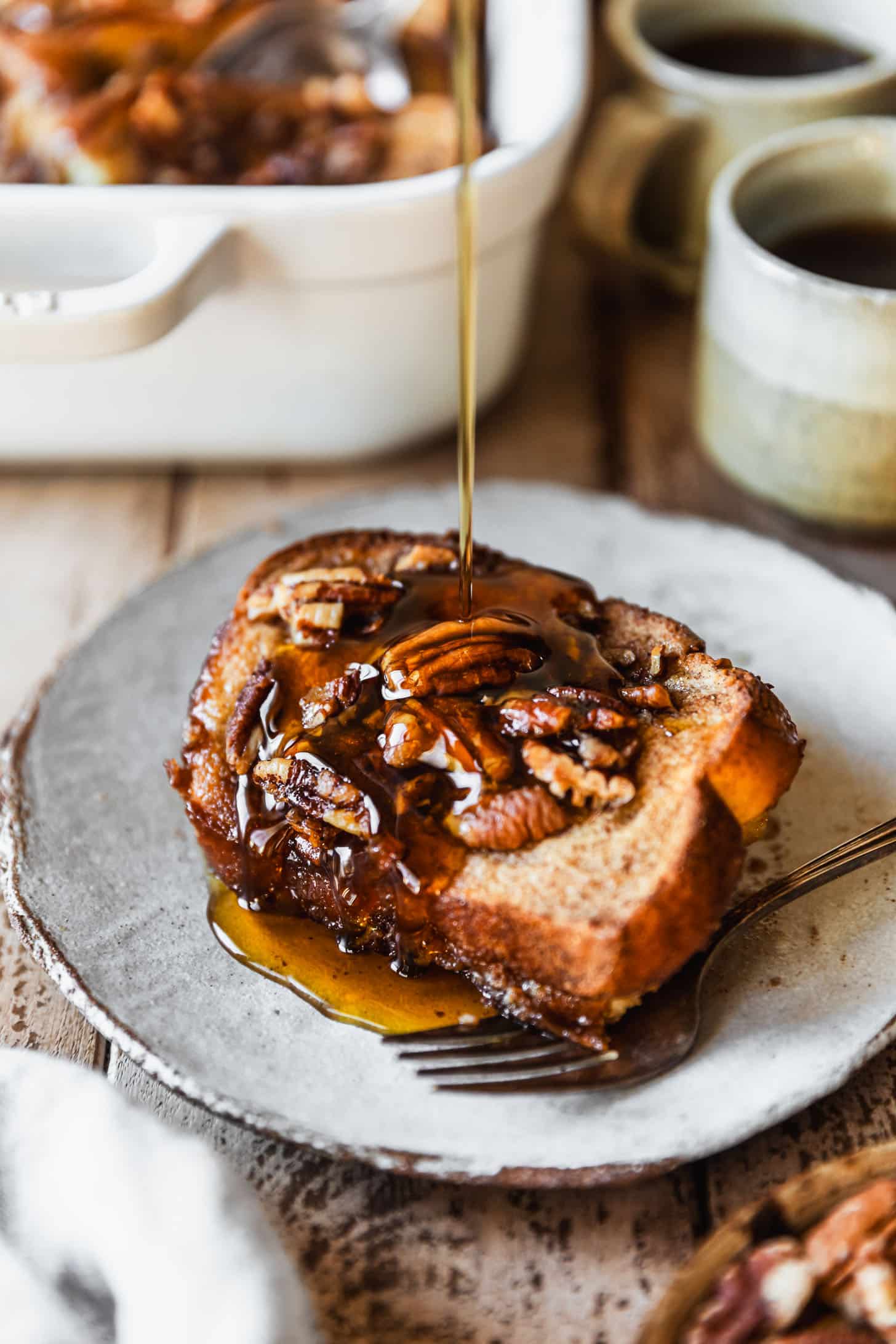 Syrup drizzling over a slice of overnight brioche French toast casserole on a grey plate next to grey cups of coffee, a white pan of casserole, and a white linen on a wood table.