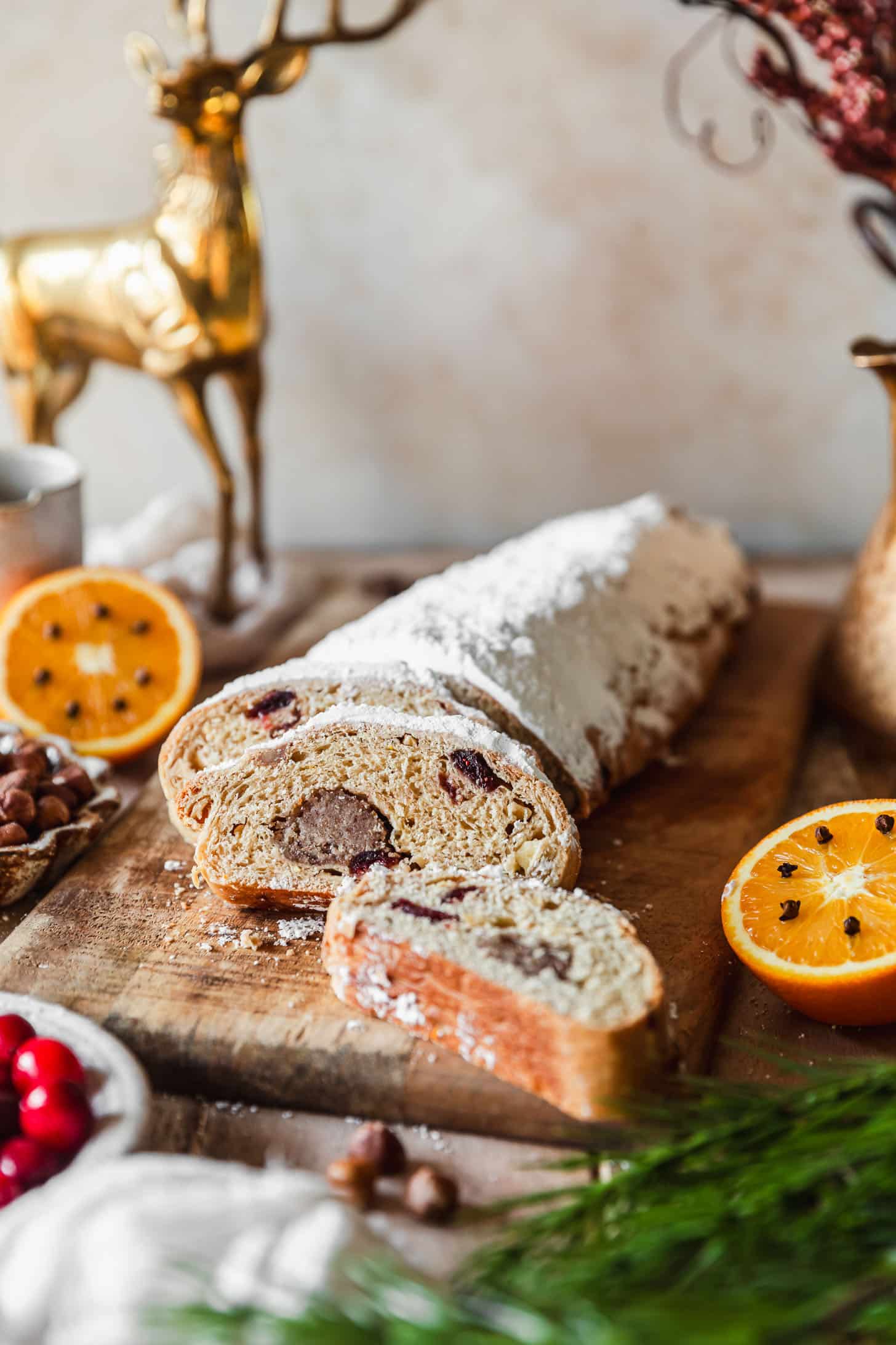 A loaf of cranberry apple stollen on a wood board next to oranges, garland, a white bowl of cranberries, and gold Christmas decorations.