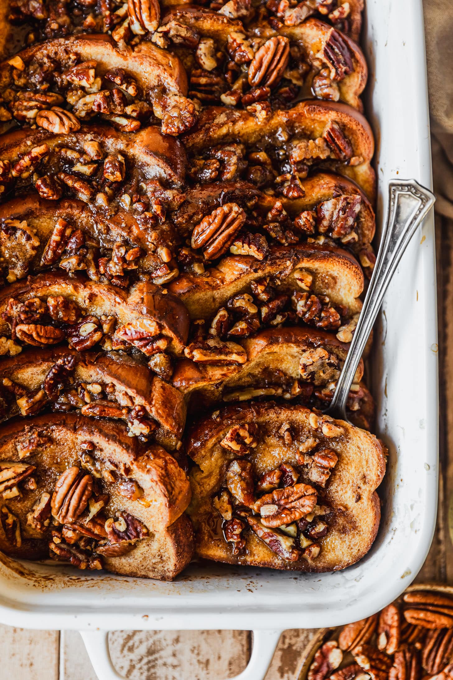 A white pan of brioche French toast casserole with pecans on a wood table next to a brown plate of pecans and a beige linen.