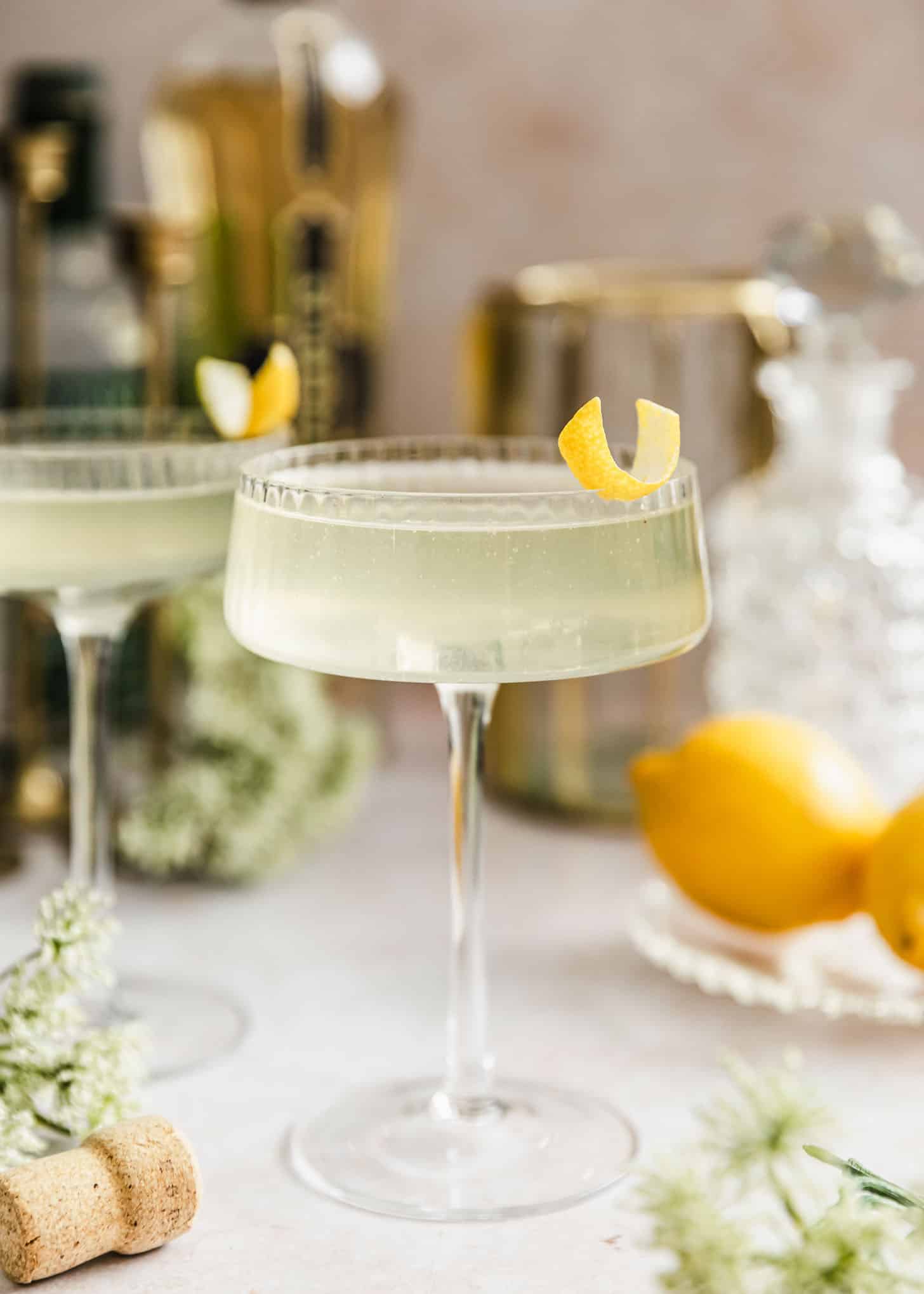 Ideas for a Cocktail Party | Two French 77 cocktails on a beige counter next to white flowers, lemons, and a bottle of elderflower liqueur.