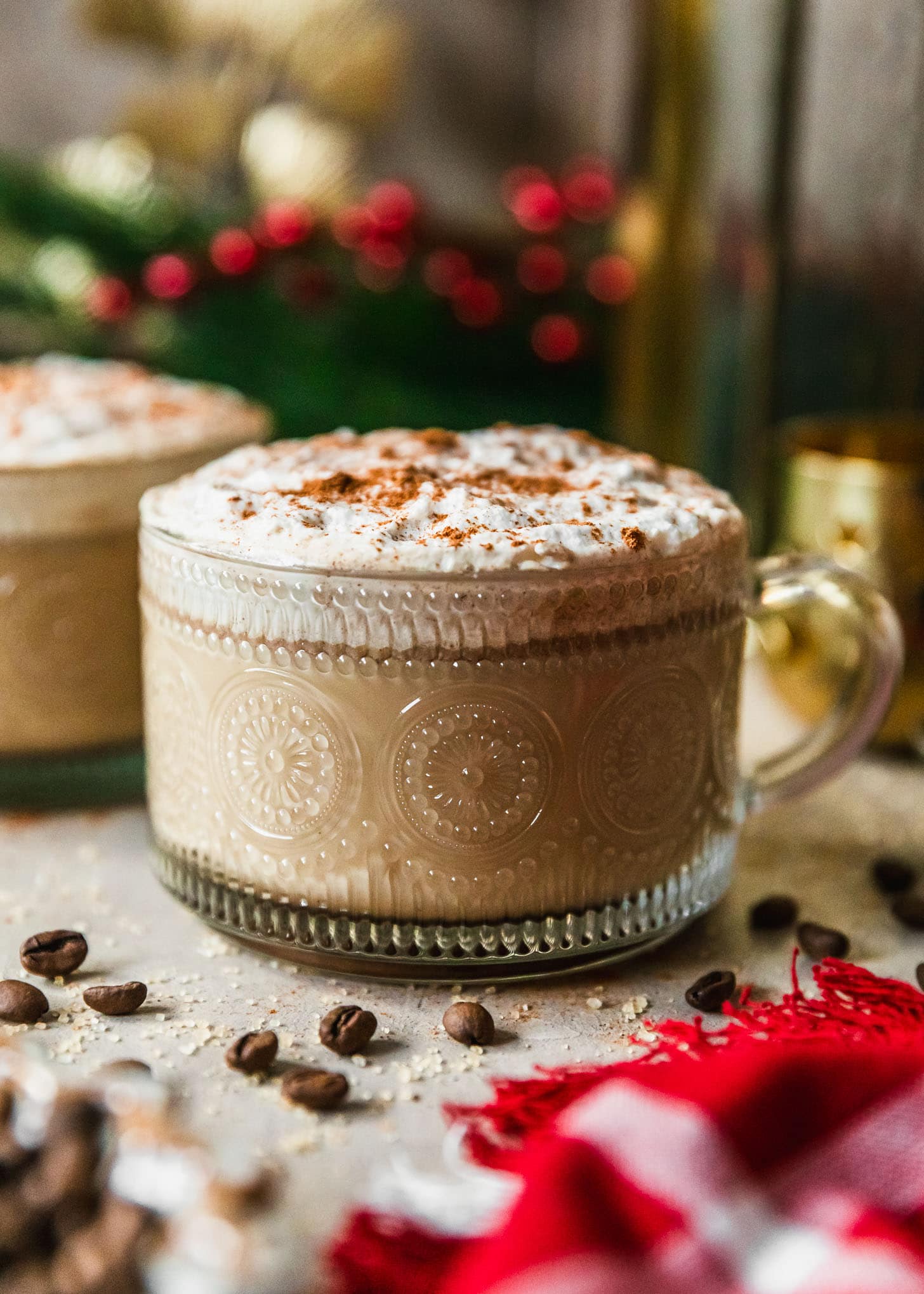 A mug of coffee hot buttered rum on a beige counter next to a red plaid linen, gold Christmas decorations, and garland.