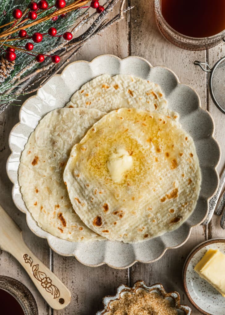 A beige plate of lefse with butter on a wood board next to glass mugs of coffee, a lefse stick, garland, and brown bowls of sugar and butter.