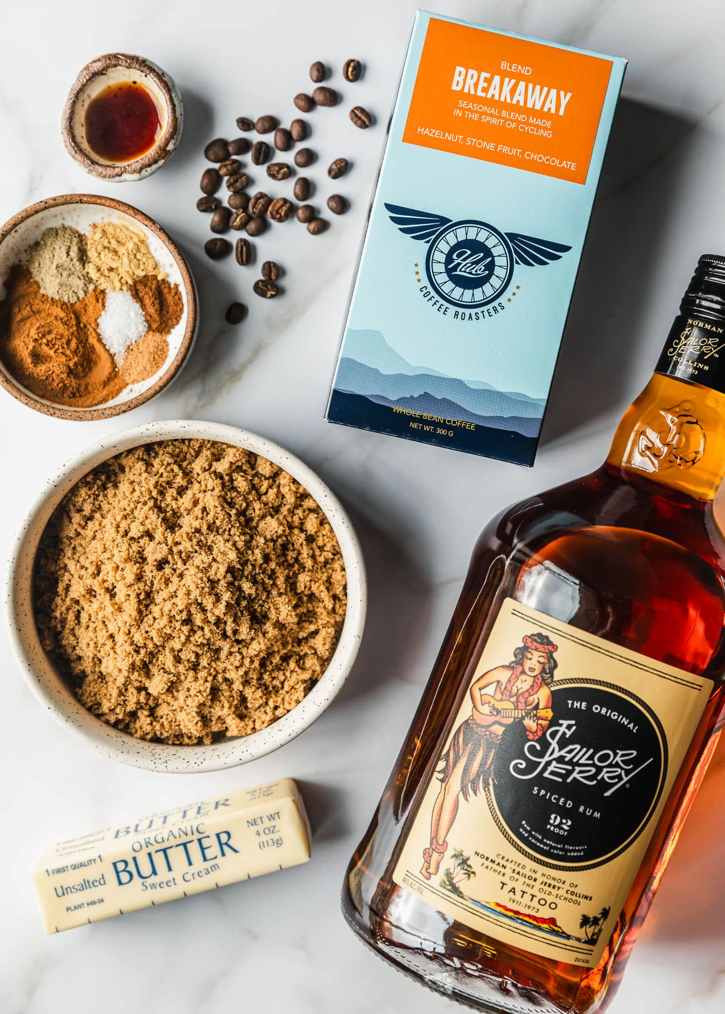 A bottle of rum, stick of butter, and box of coffee next to brown bowls of brown sugar, spices, and vanilla on a white marble counter.