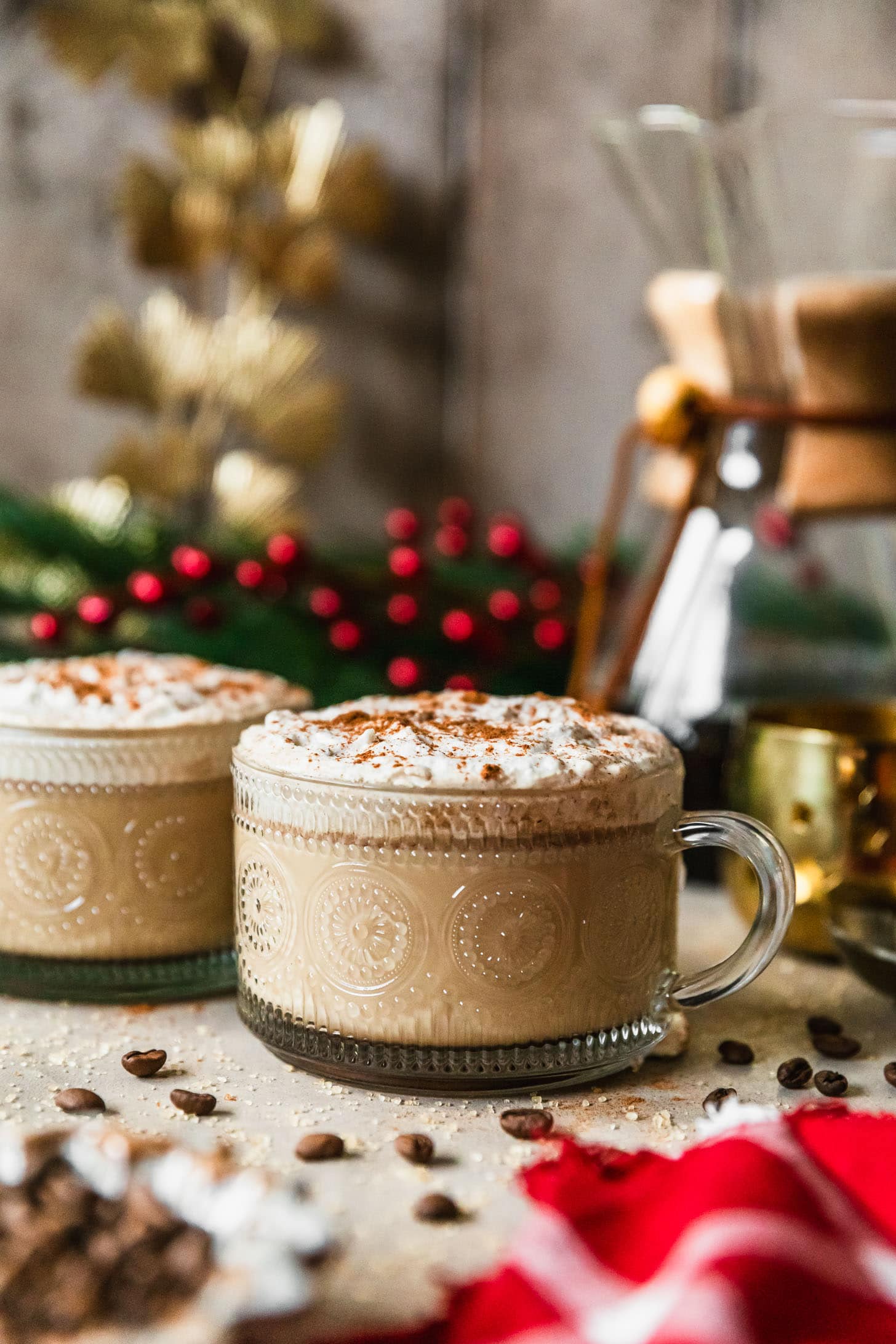 Two mugs of coffee hot buttered rum on a beige counter next to a red plaid linen, coffee beans, garland, and gold decorations.