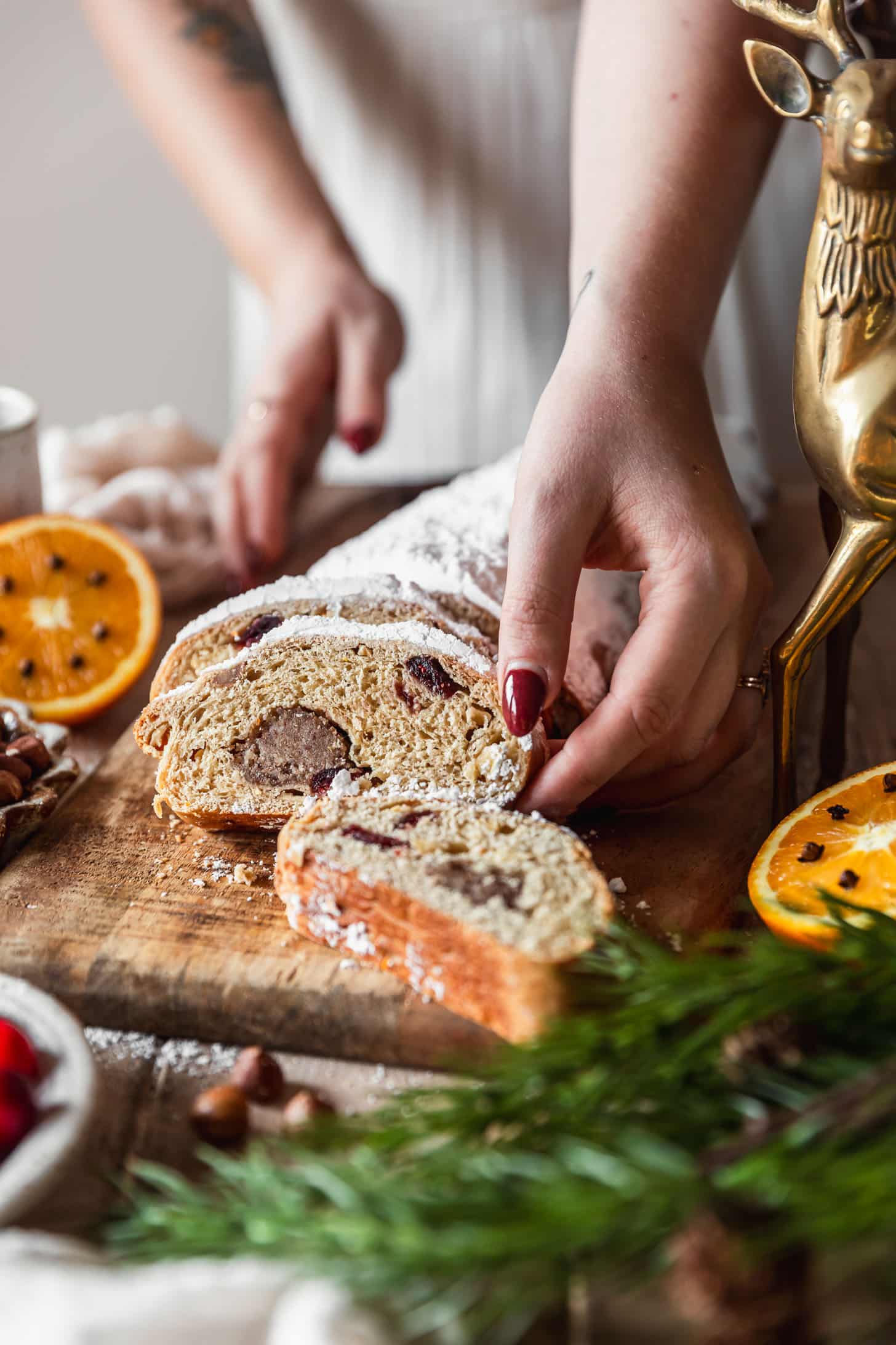 A woman in a white dress reaching for a piece of German Christmas bread on a wood board next to oranges, a gold deer, garland, and hazelnuts.