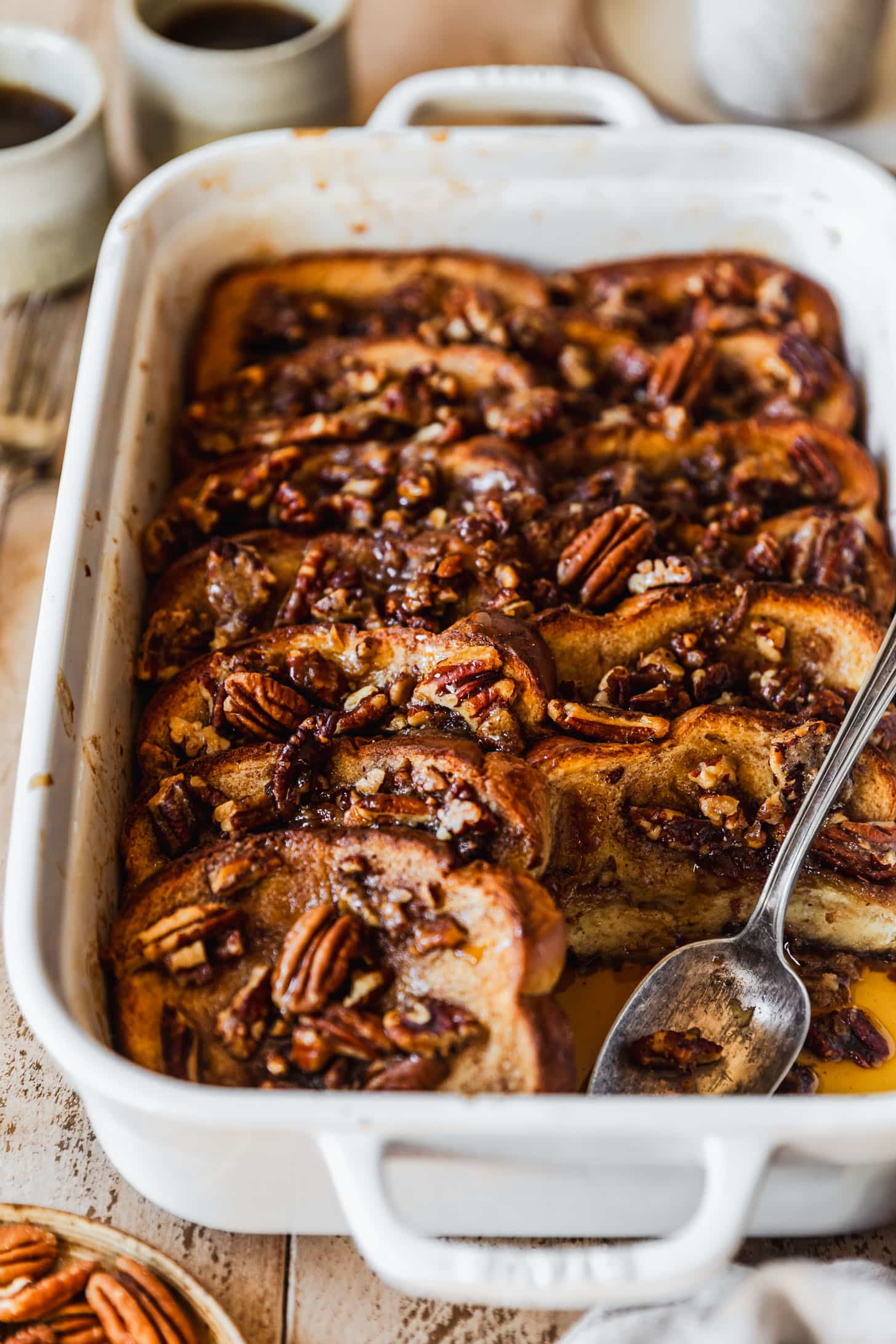 A white pan of pecan brioche French toast casserole on a wood table next to a beige linen and brown plate of pecans.