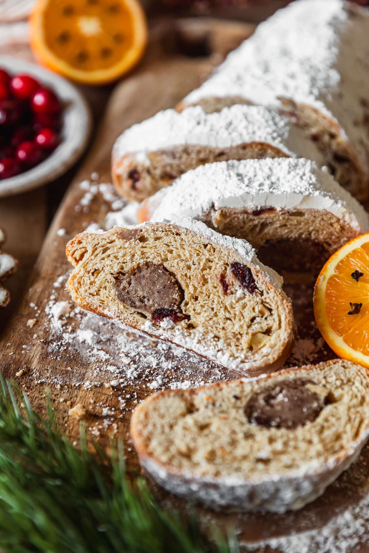 Cranberry apple stollen on a wood board next to oranges, a white bowl of cranberries, and garland on a wood table.