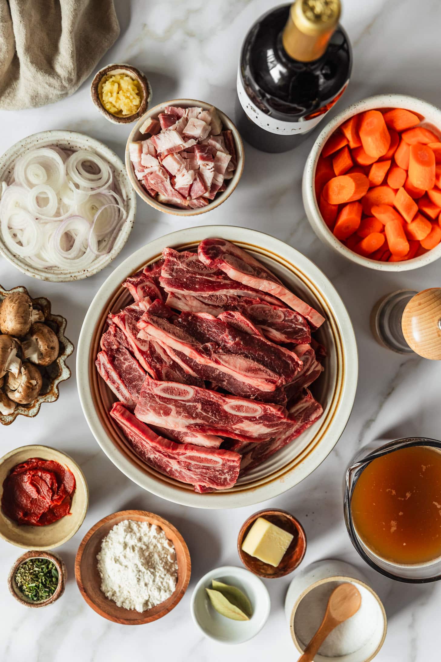 White and brown bowls of short ribs, carrots, onions, bacon, mushrooms, tomato paste, herbs, butter, and salt on a white counter next to beef broth and a bottle of wine.