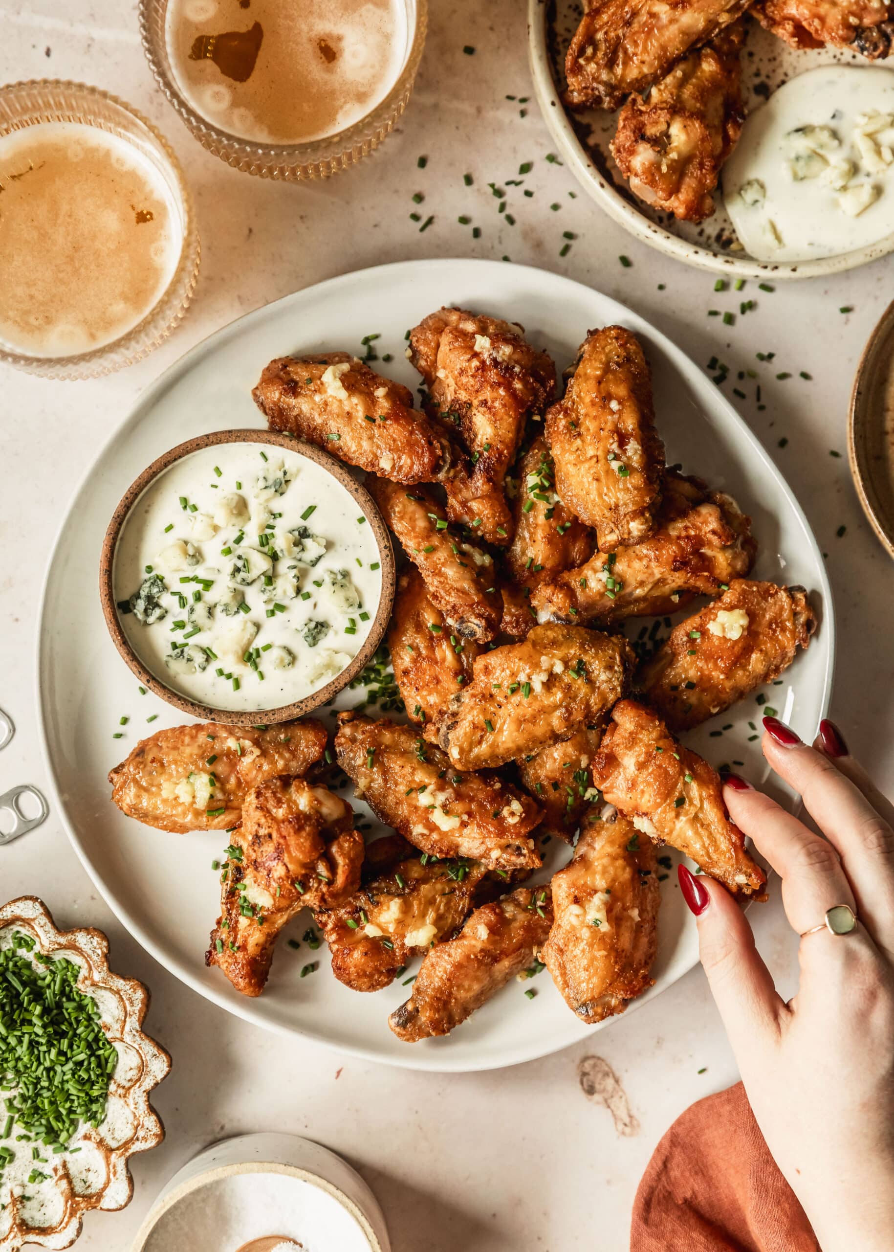 A hand reaching for a garlic butter chicken wing on a white plate next to a brown bowl of ranch, a white bowl of chives, glasses of beer, and an orange linen on a beige counter.