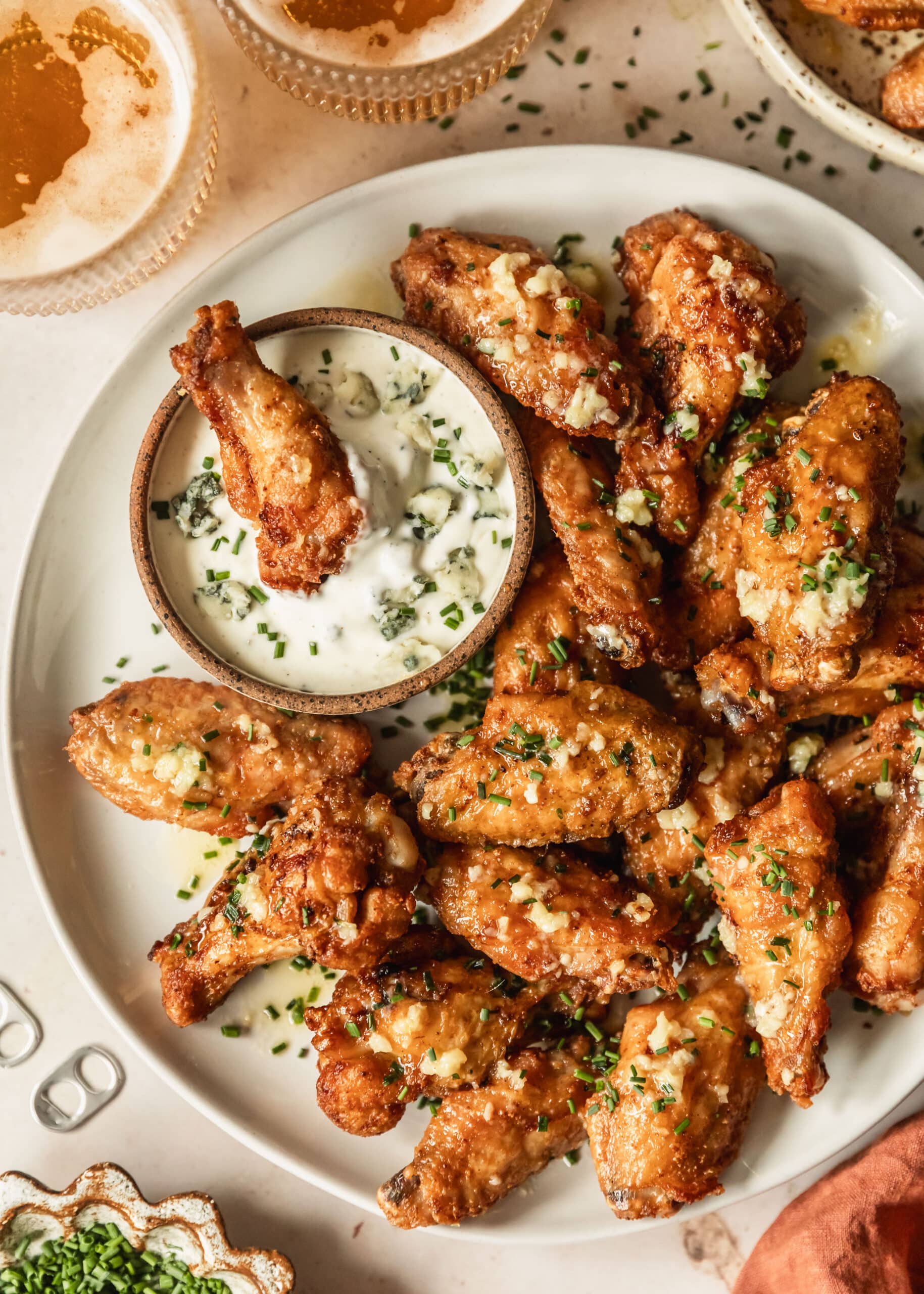 A white platter of crispy garlic butter chicken wings with a brown bowl of bleu cheese dressing on a beige counter next to beer, an orange linen, and a white bowl of chives.