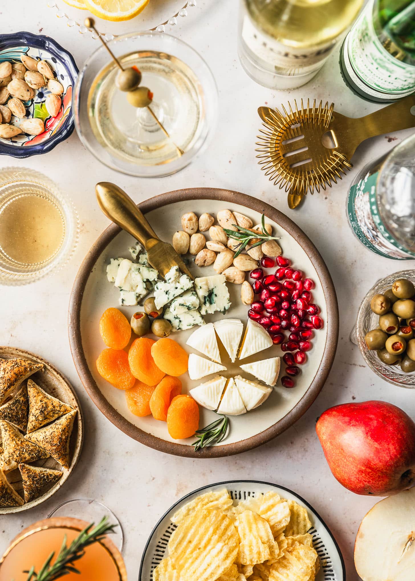 Unique Ideas for a Cocktail Party at Home | A small cheese plate, cocktails, alcohol bottles, chips, crackers, and olives on a beige counter.