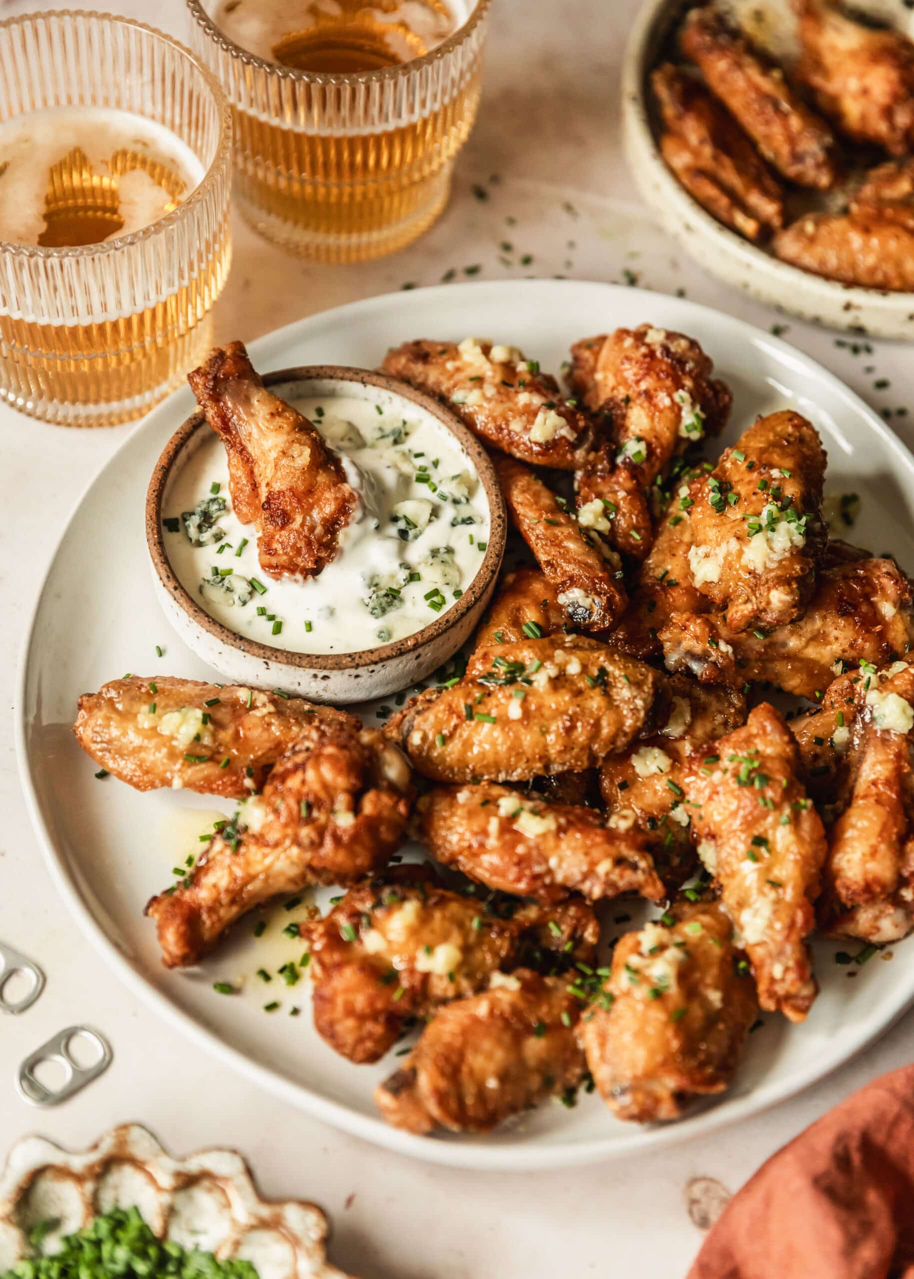 A white plate of easy garlic butter chicken wings with a white bowl of bleu cheese dressing on a beige counter next to beer, an orange linen, and a white bowl of chives.