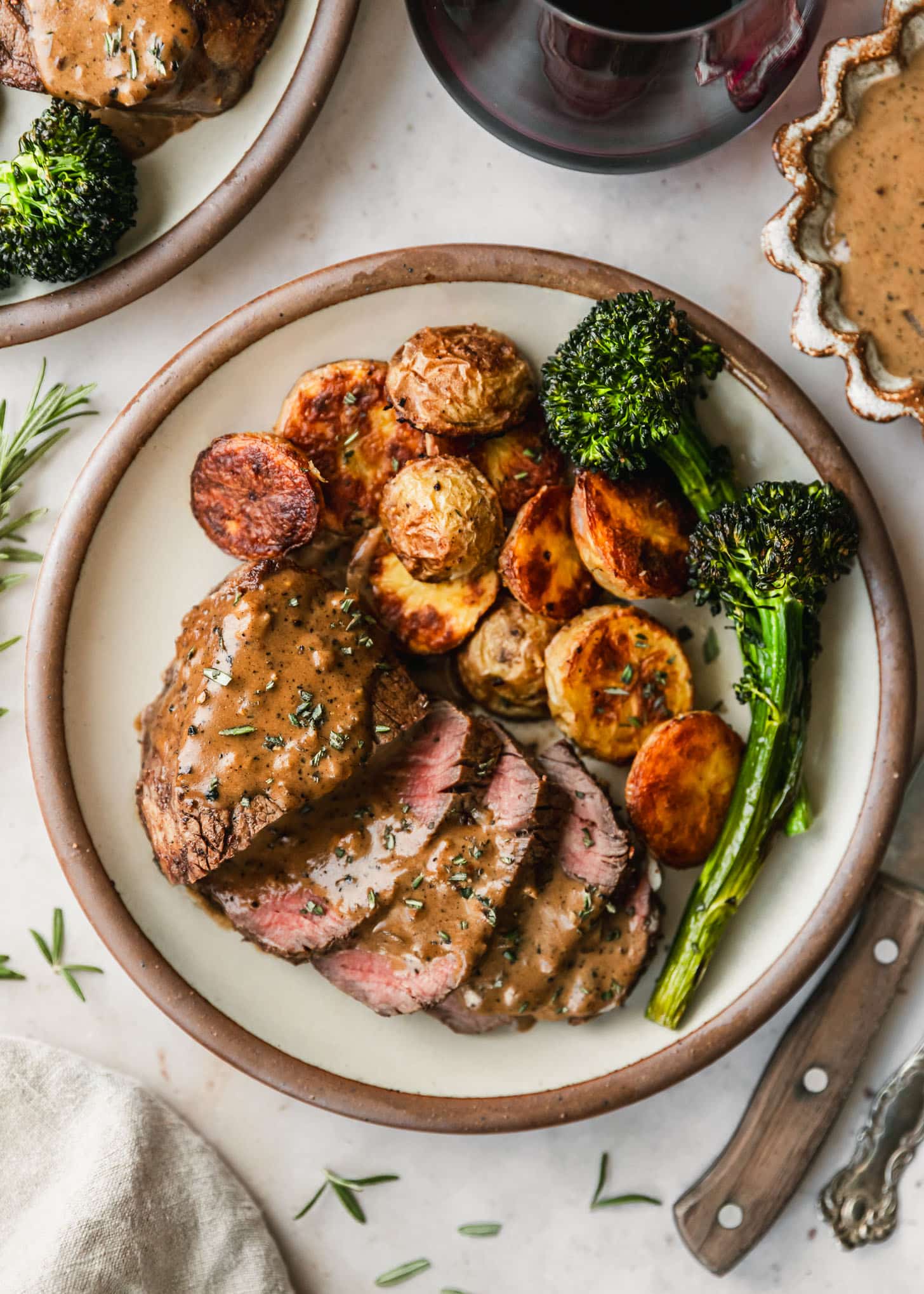Reverse sear steak, potatoes, and broccolini on a white plate next to red wine, a brown bowl of peppercorn sauce, and a wood steak knife on a marble counter.
