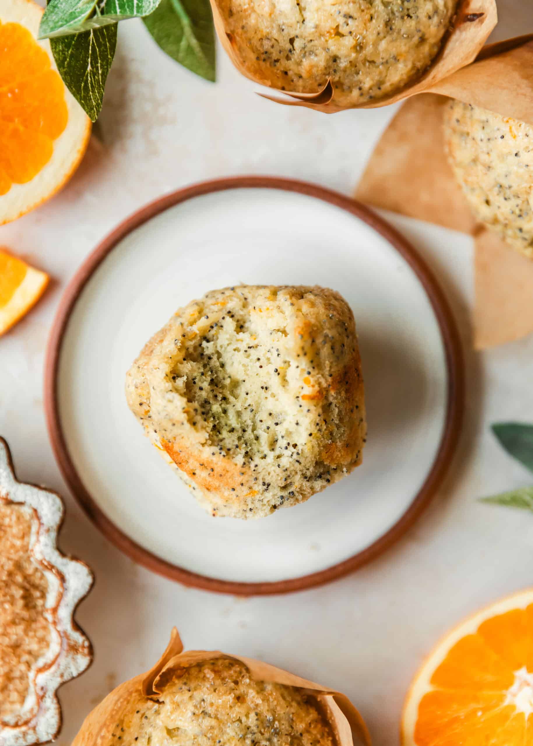 A citrus muffin with a bite taken out of it on a white plate next to sliced navel oranges, leaves, muffins, and a brown bowl of sugar on a beige counter.