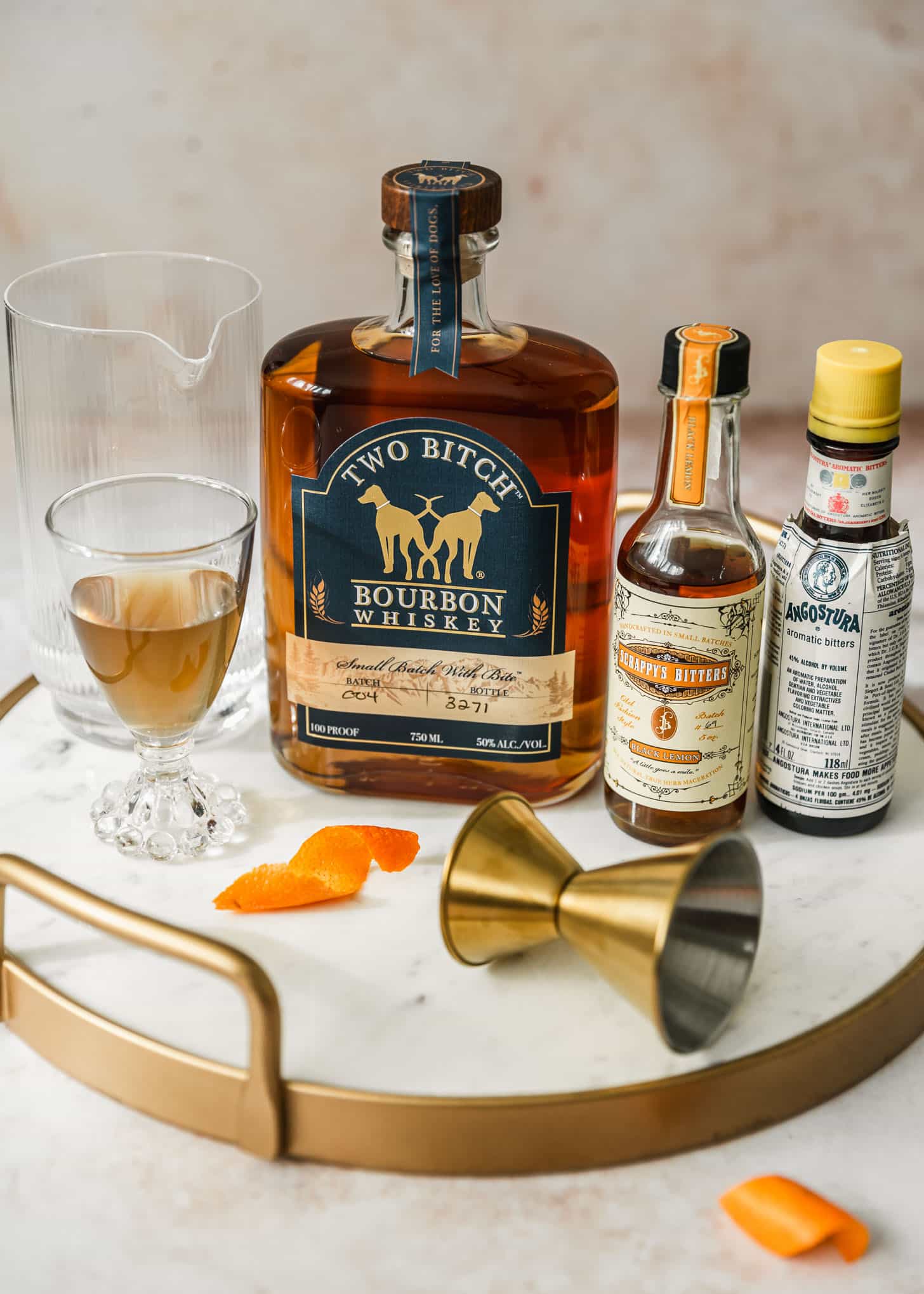 A bottle of bourbon, two bottles of bitters, mixing glass with water, cup of simple syrup, and orange peel on a marble tray with a tan background.