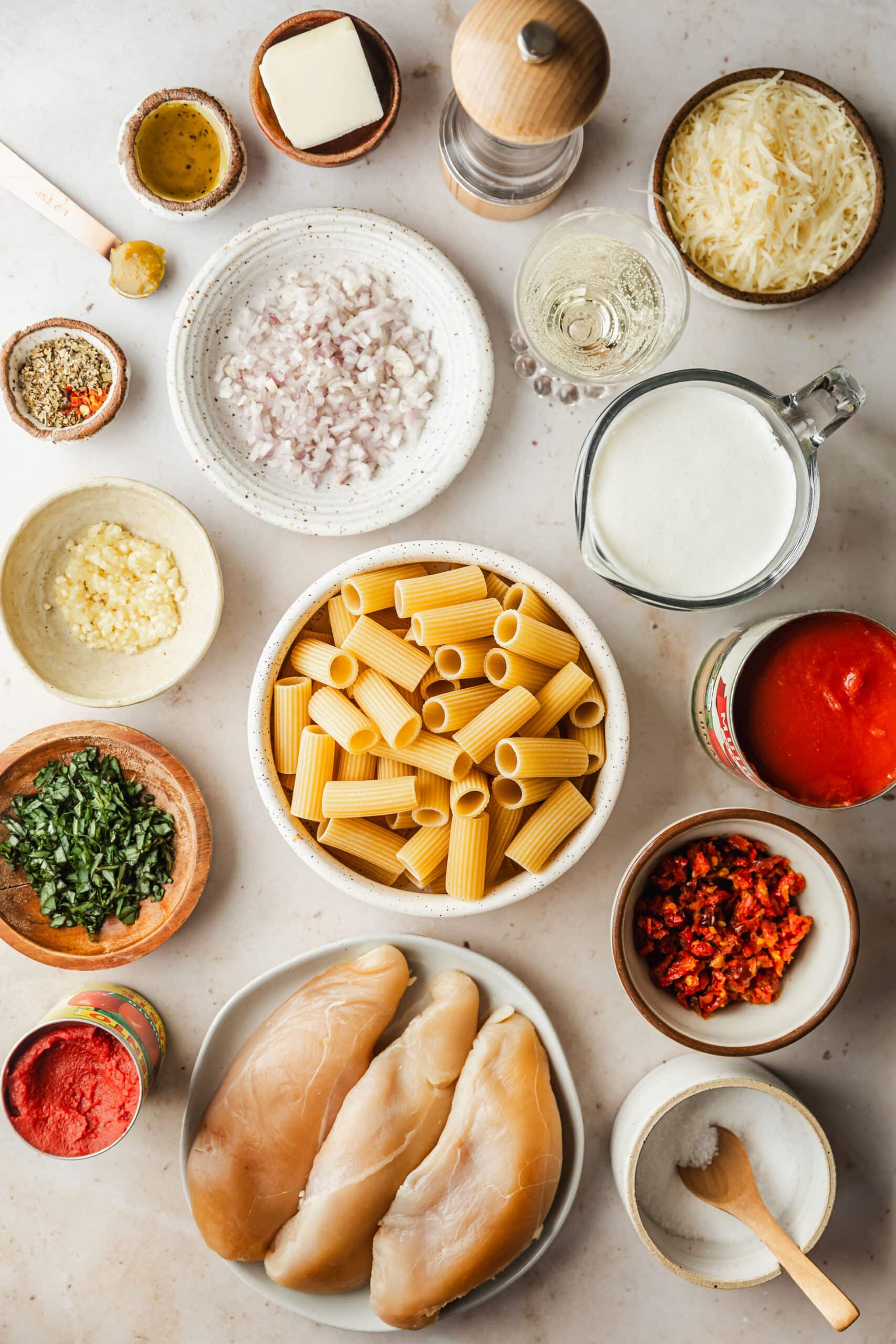 White and brown bowls of pasta, chicken, garlic, basil, salt, sundried tomatoes, cream, shallots, spices, parmesan, and white wine next to cans of tomatoes on a beige counter.