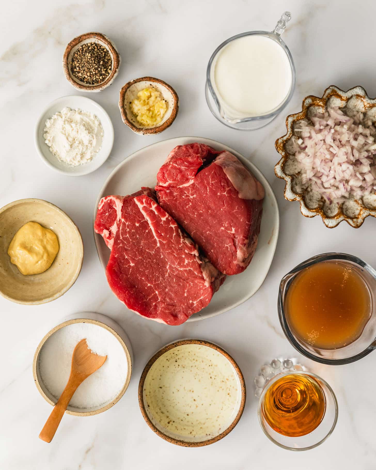 White and brown plates of steak, shallots, cream, garlic, pepper, flour, mustard, salt, oil, brandy, and beef broth on a beige marble counter.