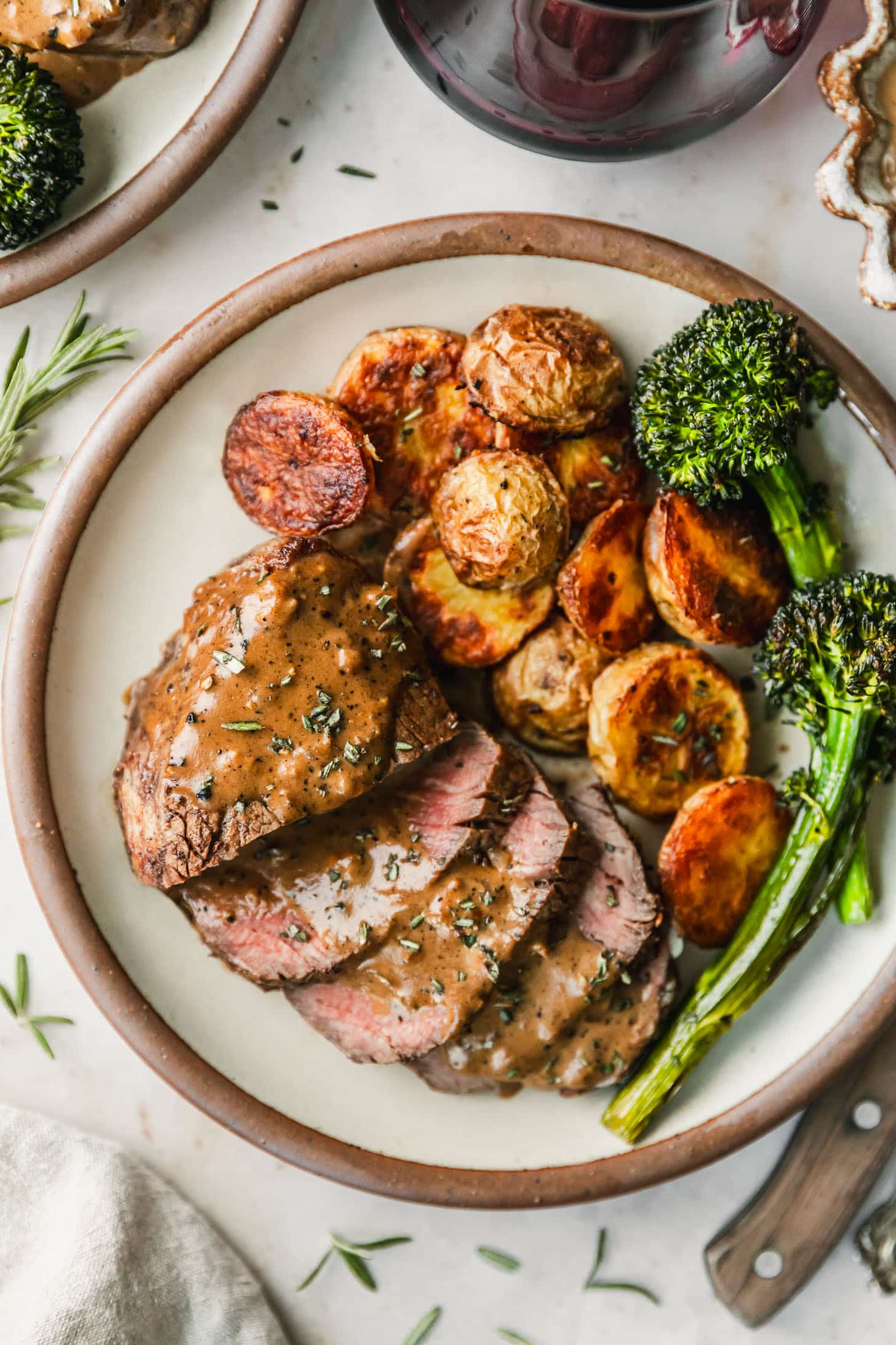 A white plate with potatoes, broccolini, and reverse sear filet mignon with peppercorn sauce on a beige marble counter next to a wood knife, beige linen, and red wine.