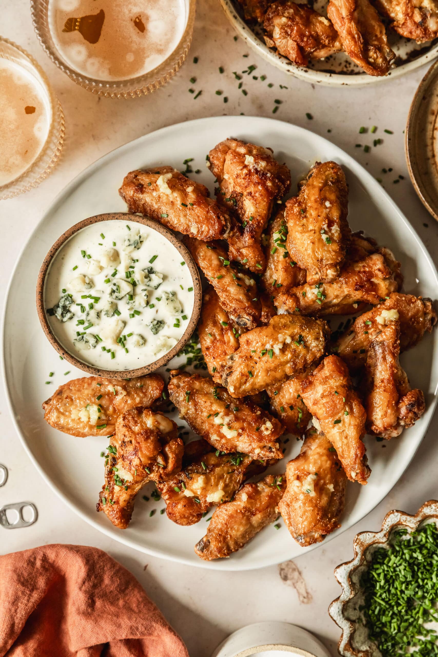 A white platter of garlic butter chicken wings and a brown bowl of ranch on a beige counter next to glasses of beer, an orange linen, and a white bowl of chives.