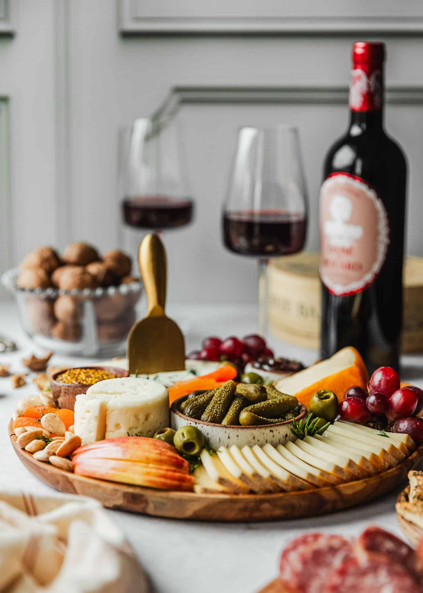A French cheese board on a white counter next to red wine, a bowl of walnuts, a beige linen, and crackers.