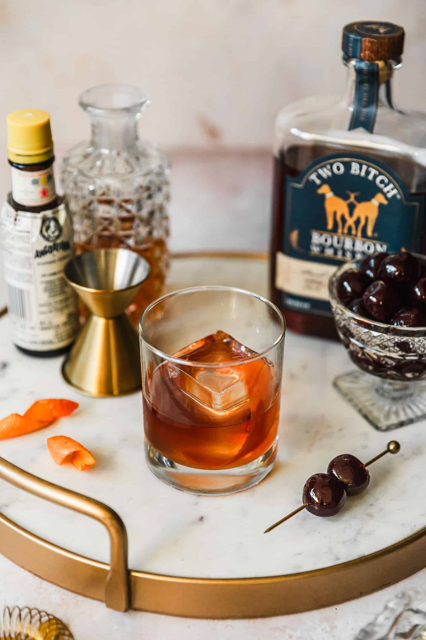 A freezer door old fashioned on a marble tray next to bourbon, bitters, a gold shot glass, and a glass bowl of cherries with a beige background.